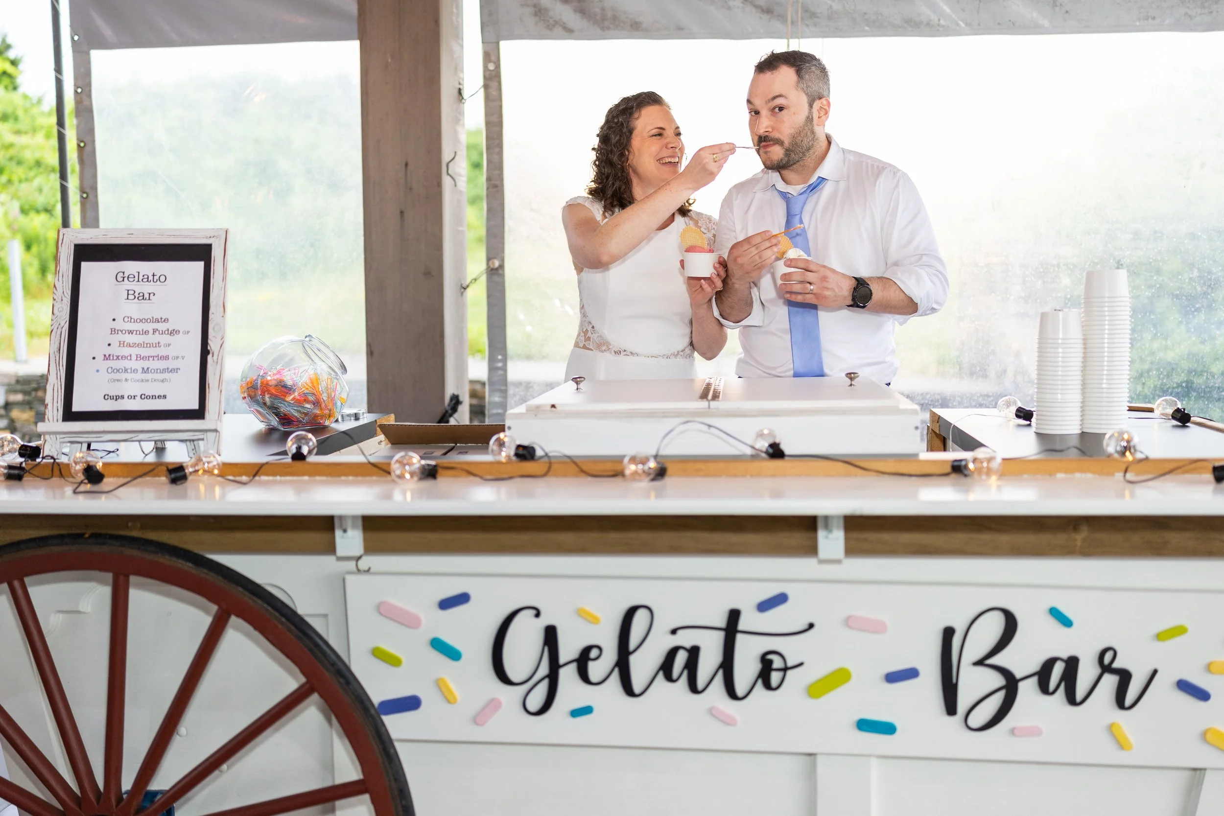 A woman feeding a man with a toothpick at a gelato bar. The bar has a sign listing gelato flavors like chocolate, brownie fudge, hazelnut, mixed berries, and cookie monster. The woman is smiling and holding a cup of gelato, while the man is holding a