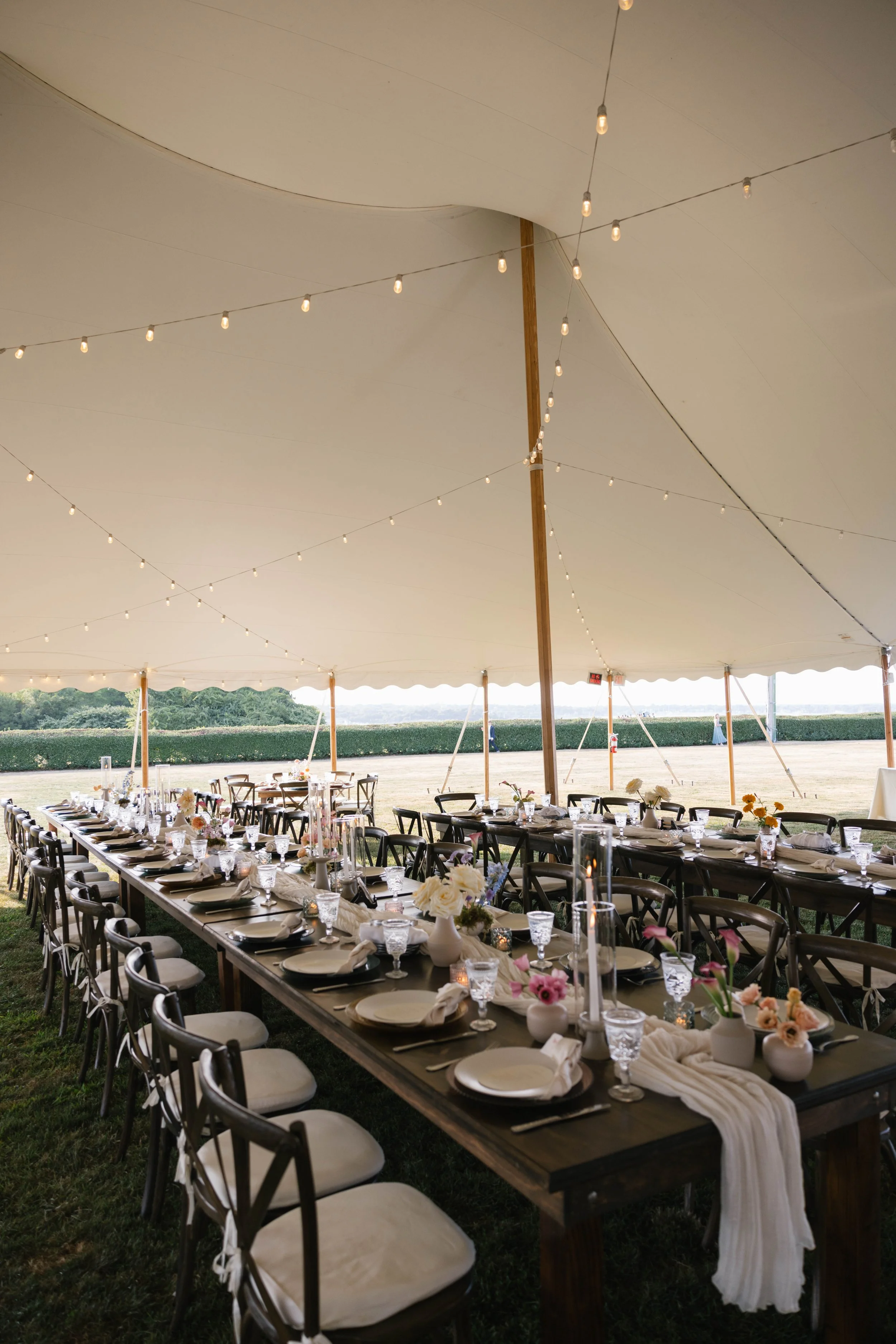 Long dining tables decorated for an outdoor event, under a large tent with string lights hanging from the ceiling. The tables are set with white plates, glasses, napkins, and floral centerpieces, with chairs lined along the sides of the tables. In th