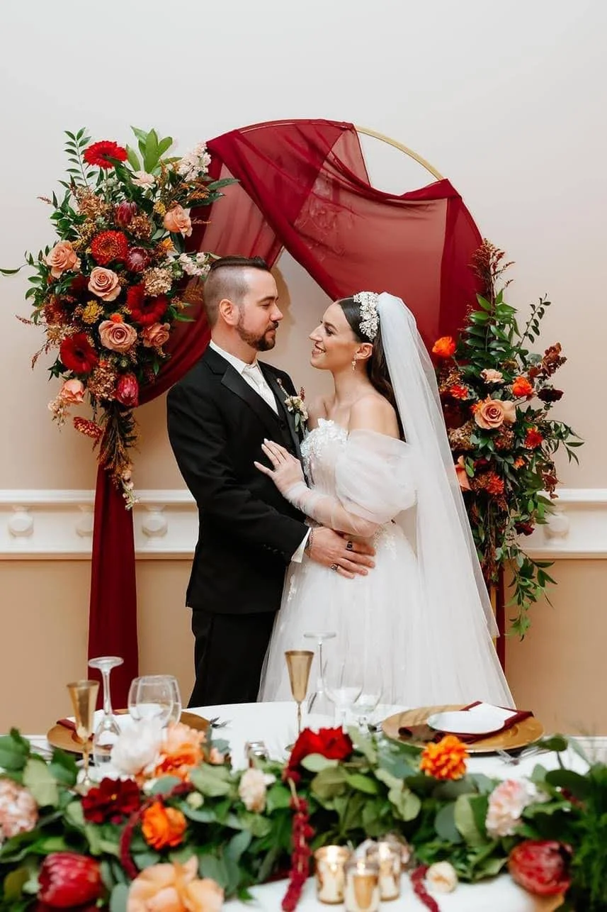 A bride and groom standing together at their wedding, looking at each other. The bride is wearing a white wedding dress with lace details and a veil, while the groom is in a black suit and white shirt. They are in front of a floral backdrop with red 