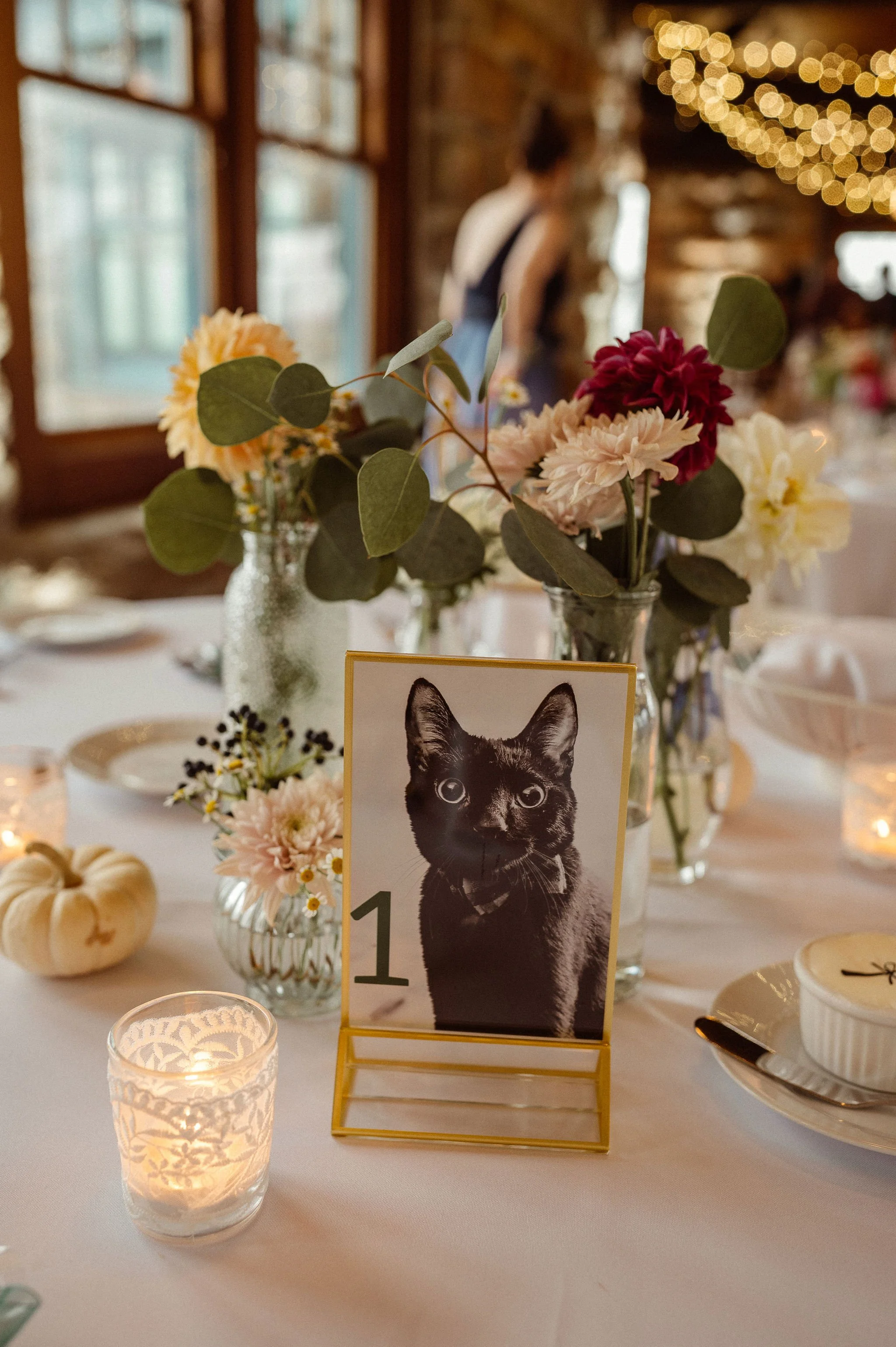 Table decorated for a celebration with floral arrangements, candles, small pumpkins, and a photo of a black cat with large eyes.