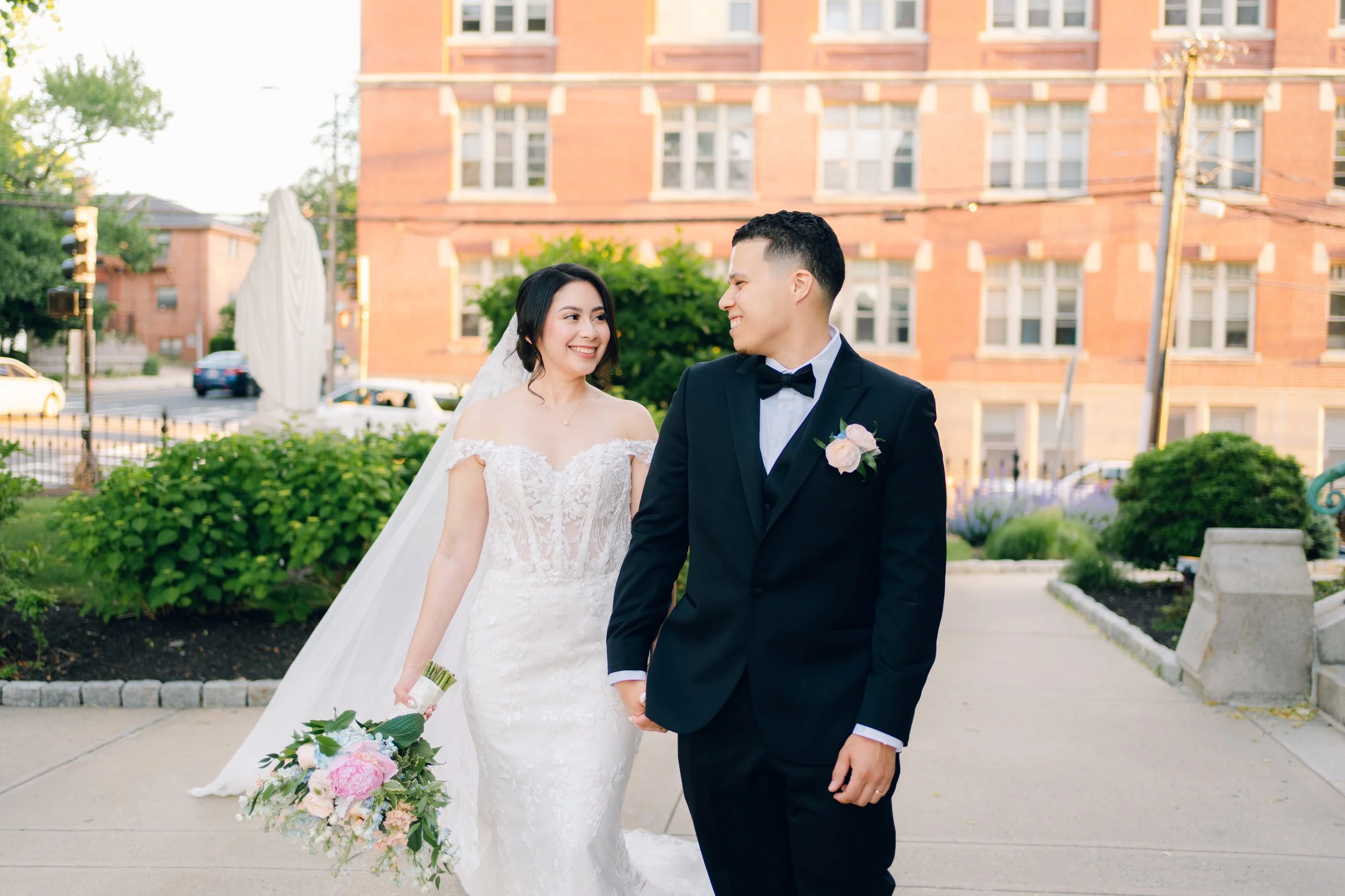 A bride and groom holding hands and smiling at each other outdoors in front of a red brick building, with greenery and a bouquet of flowers.