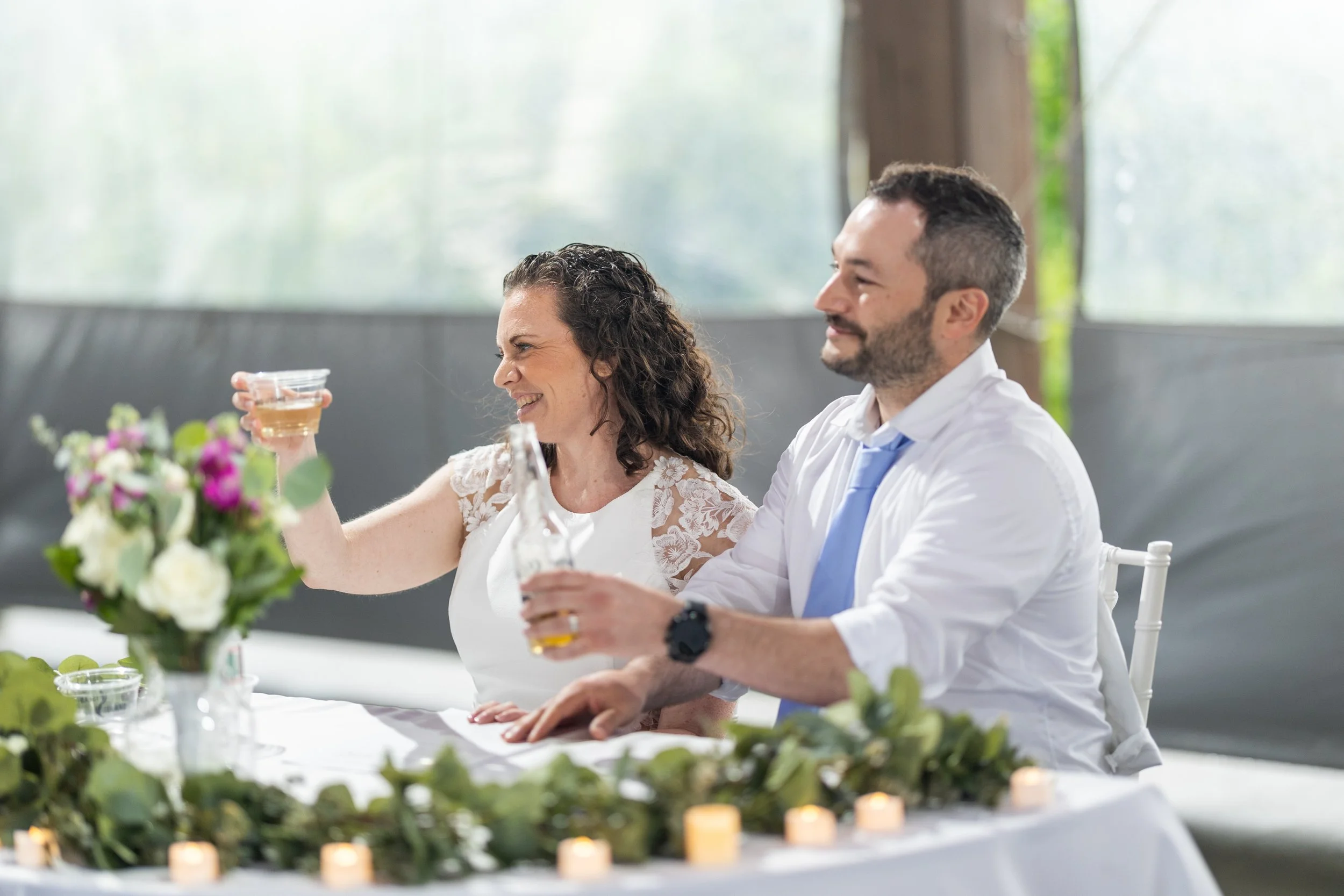 A woman and a man sitting at a decorated table, smiling, with the woman holding a drink. There are flowers and candles on the table, and a window with greenery outside in the background.