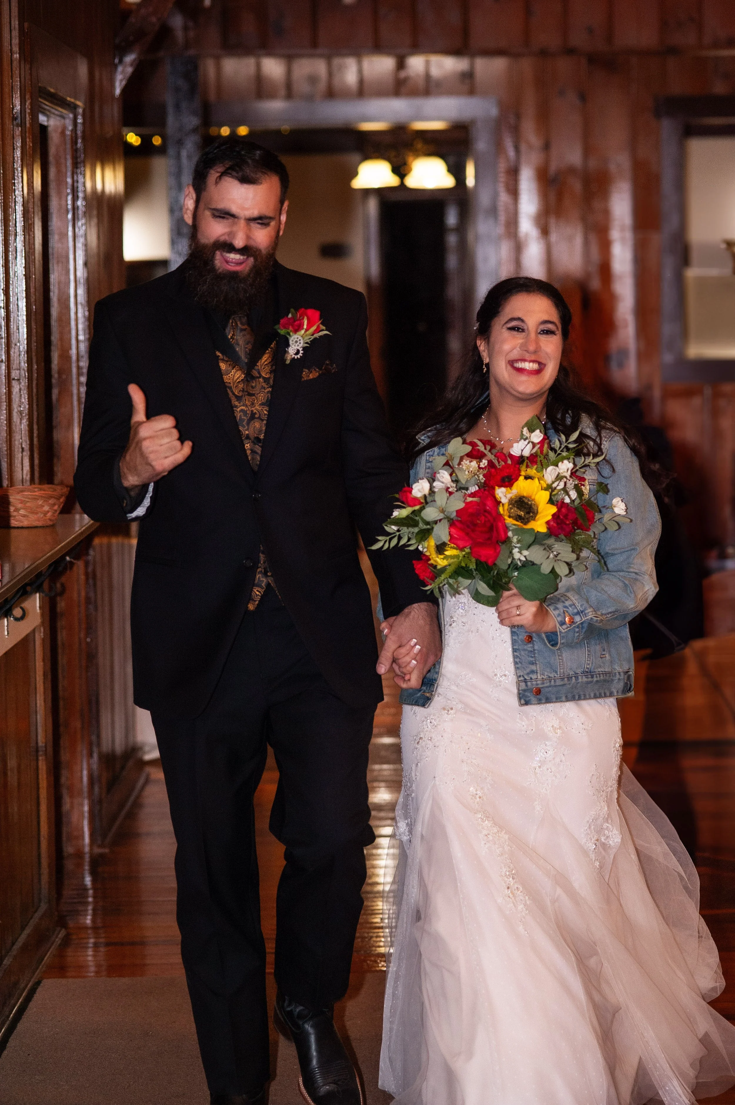 A man and woman dressed in wedding attire holding hands and smiling in a wooden interior space, with the woman holding a colorful bouquet.