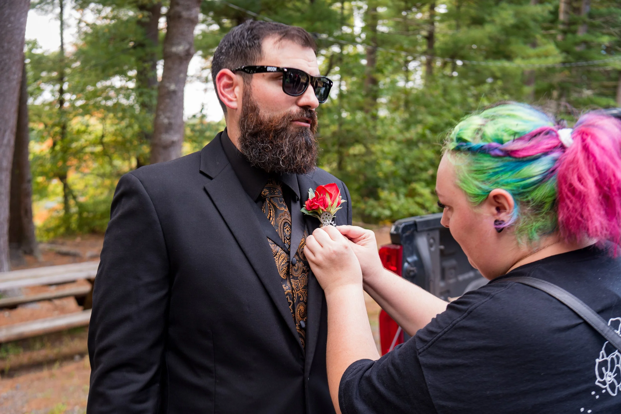 A man in sunglasses and a suit is receiving a flower boutonniere from a woman with colorful rainbow hair outdoors with trees in the background.
