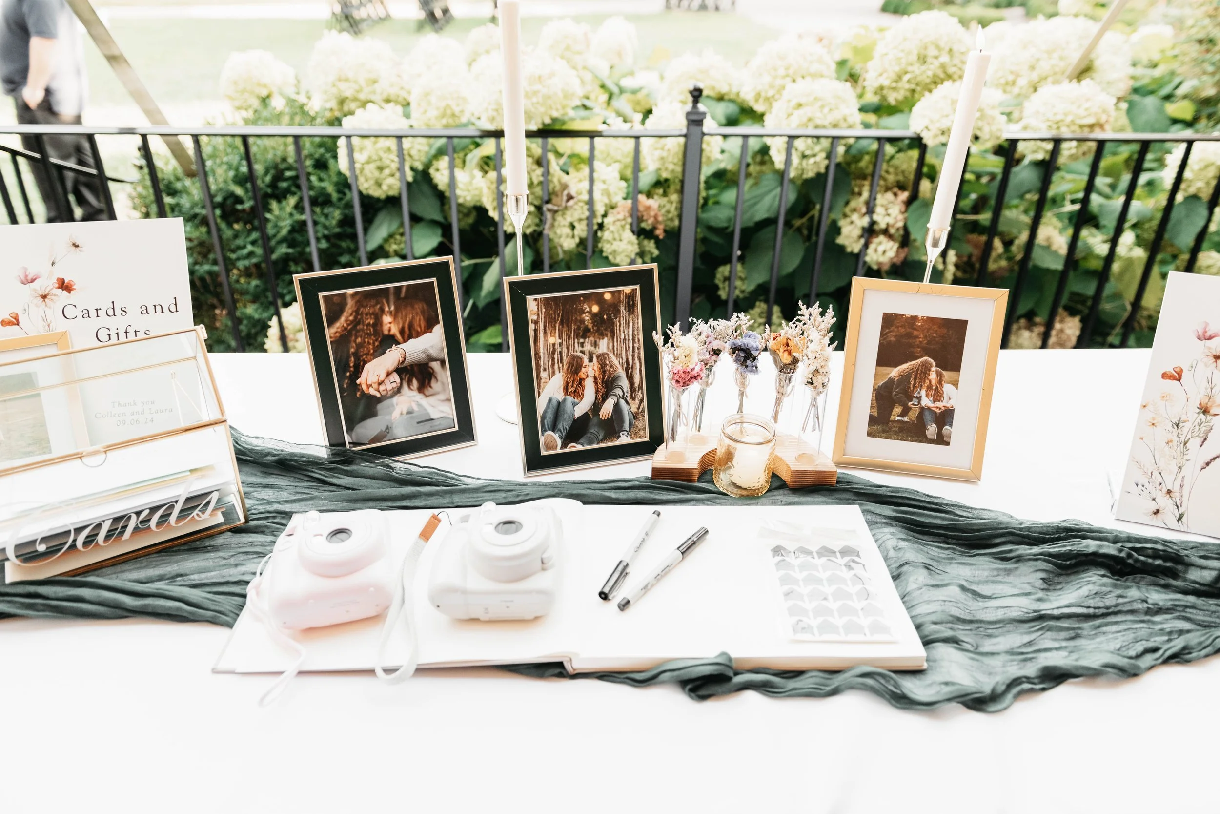 Display of framed photographs, floral arrangements, candles, and greeting cards at an outdoor event space with green plants and hydrangeas in the background.