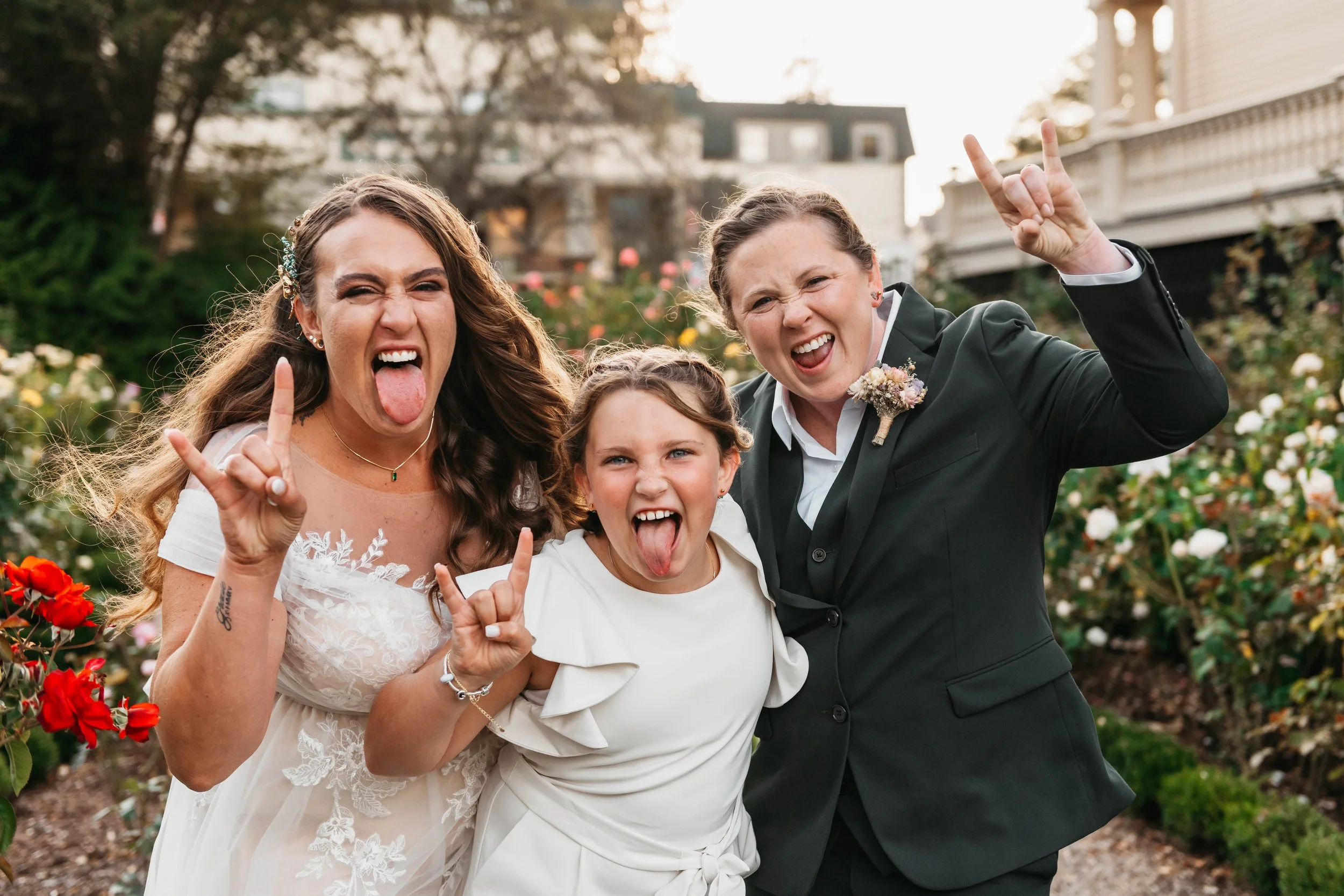Three women, two adults and a child, making rock on hand gestures, sticking out their tongues, and smiling in a garden with flowering bushes and trees in the background during sunset.