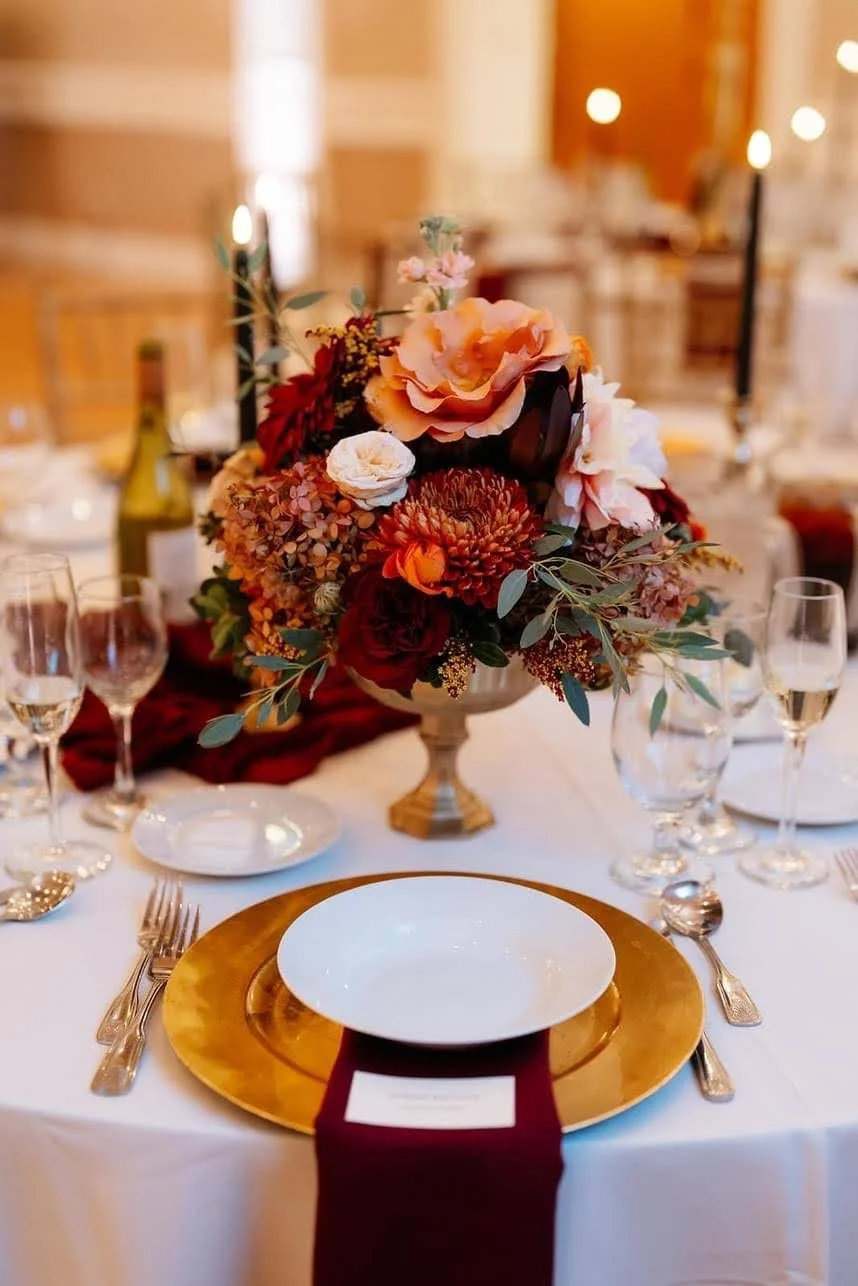 Elegant table setting with a large floral centerpiece featuring pink, orange, and red flowers, around which there are plates, silverware, wine glasses, and a chair with a white cushion, set in a warmly lit room.