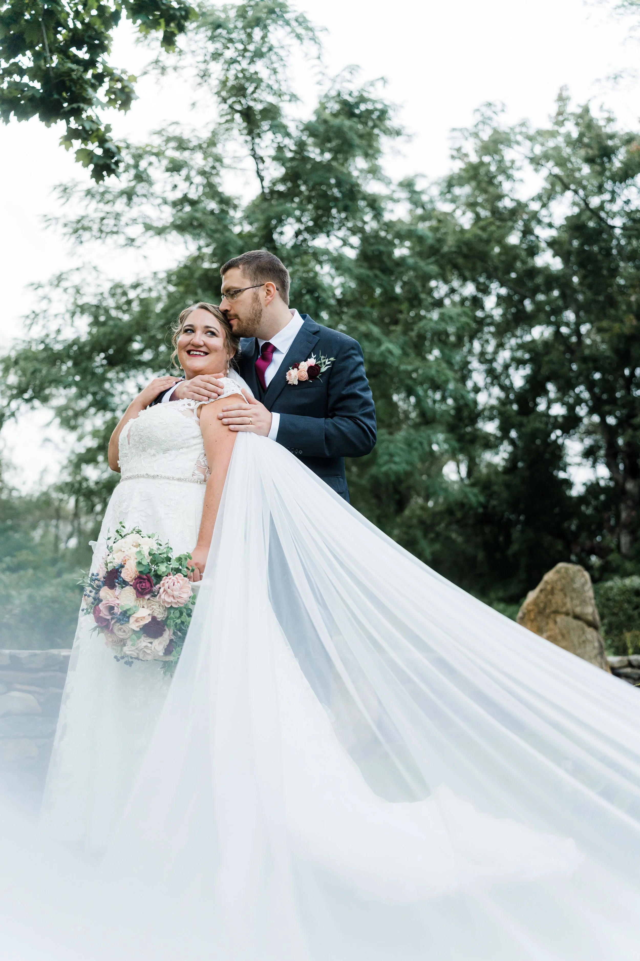 A bride and groom embracing outdoors, with the bride holding a floral bouquet, surrounded by green trees, during a wedding photoshoot.