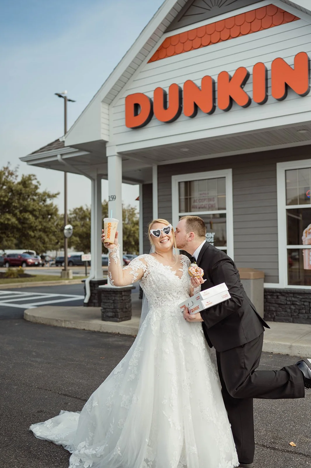 A bride and groom in wedding attire enjoying their time at a Dunkin' Doughnuts restaurant. The bride is smiling with sunglasses, holding a drink and a donut, while the groom is kissing her cheek and holding a box of donuts outside the store.