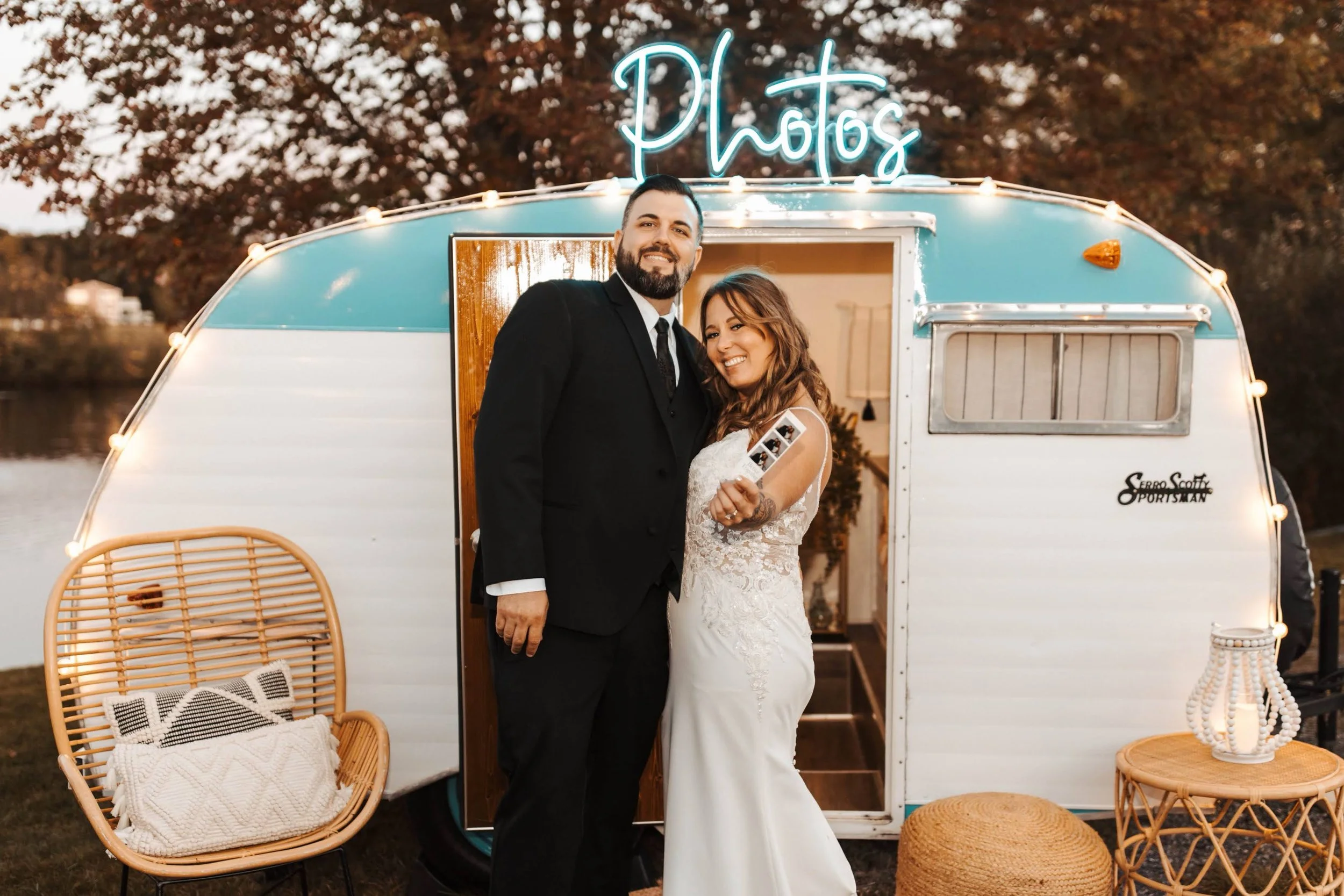 A happy couple, dressed in wedding attire, poses in front of a vintage camper with a neon sign that says 'Photos' on top. They are smiling and holding photo strips, with outdoor chairs and decor around them.