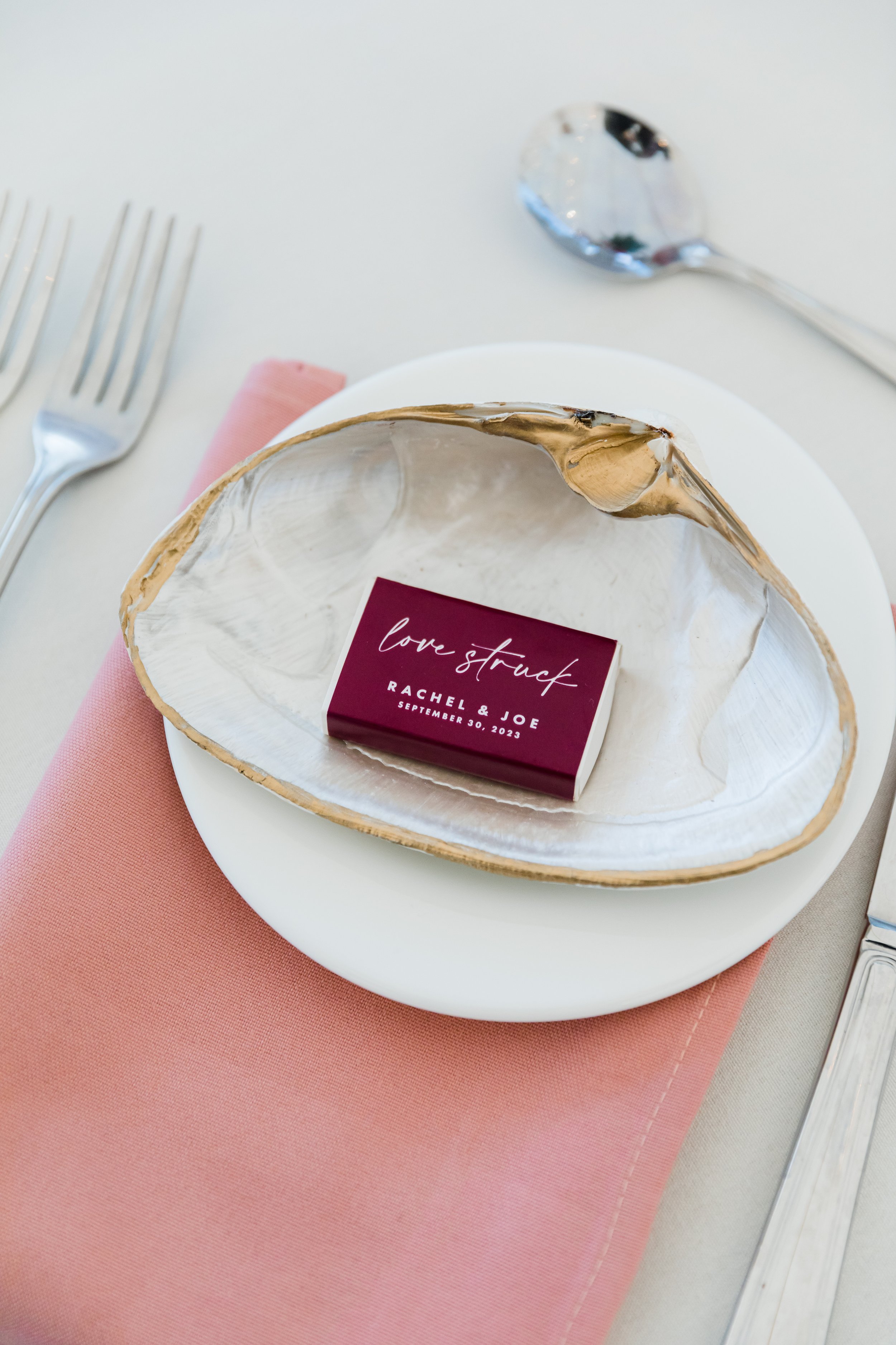 A white plate with a halved oyster shell in the center, containing a red box labeled 'love strucch' with the names 'Rachel & Joe' and a date 'September 30, 2023.' The plate is on a pink napkin, with a fork and spoon arranged on a white table.