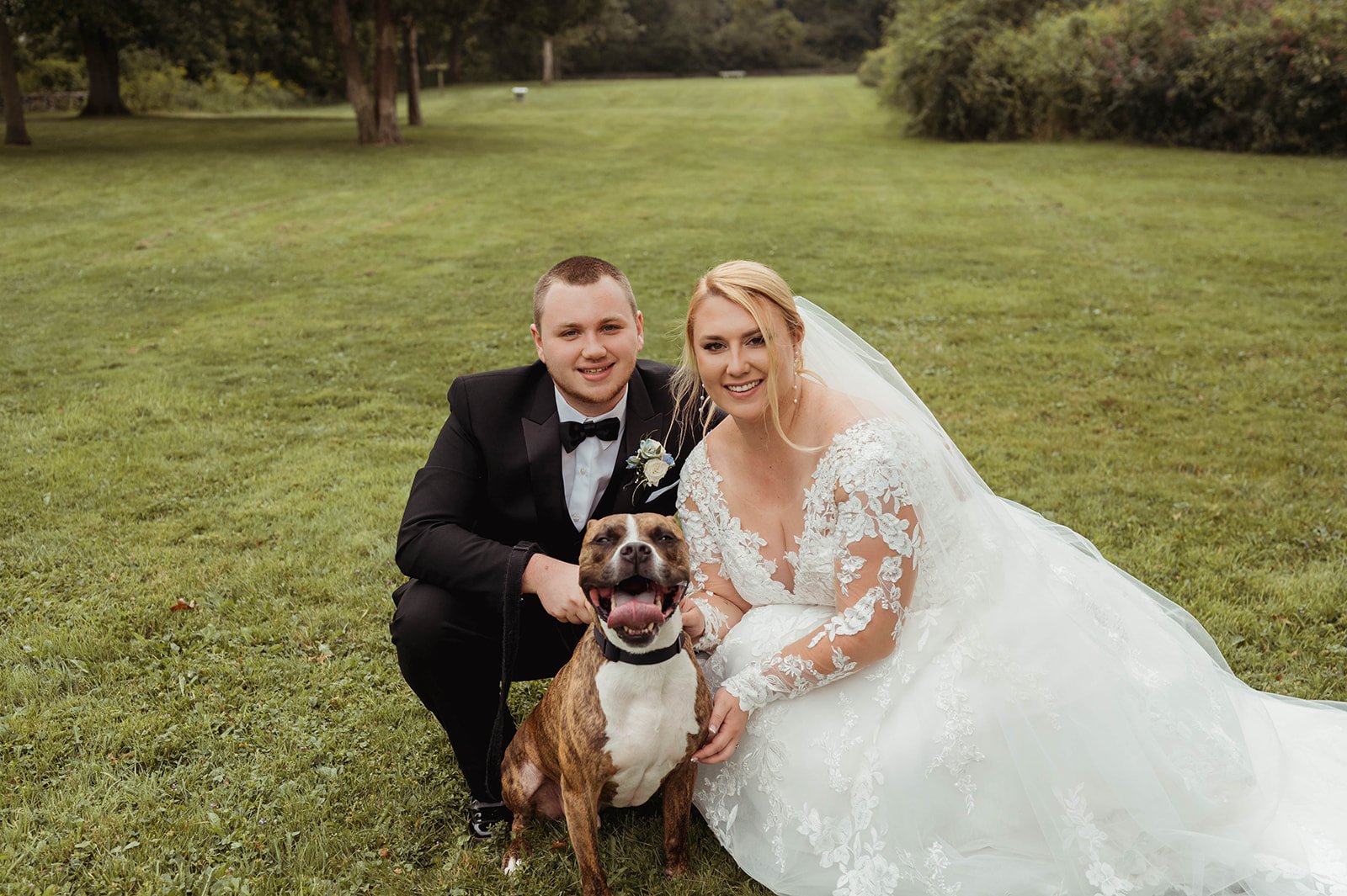 A newlywed couple in wedding attire posing with their dog on a grassy field with trees in the background.