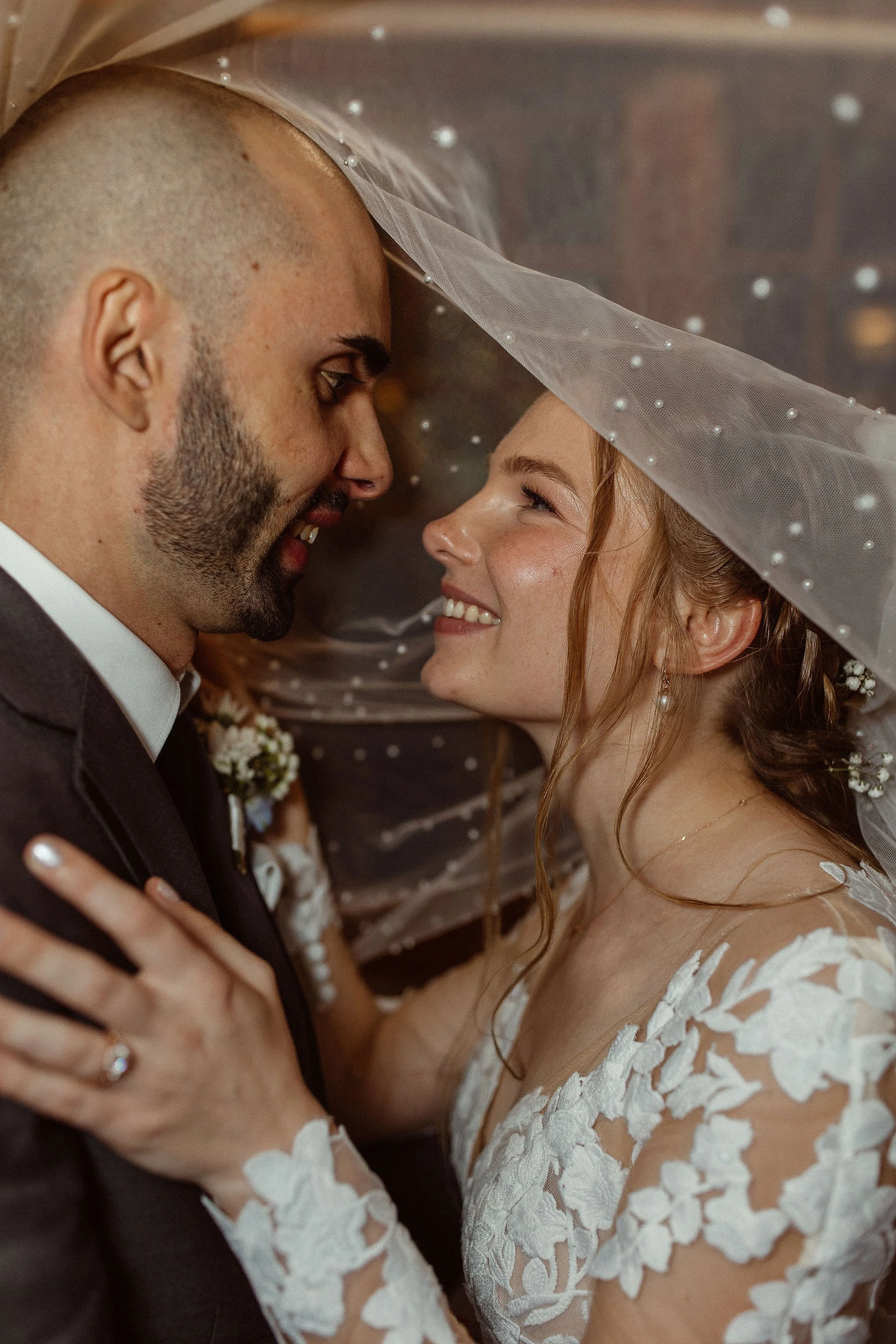 A couple on their wedding day, smiling and leaning in close under a veil with pearls. The bride has red hair and wears a white dress with lace details. The groom has a beard and bald head, wearing a dark suit with a white shirt.
