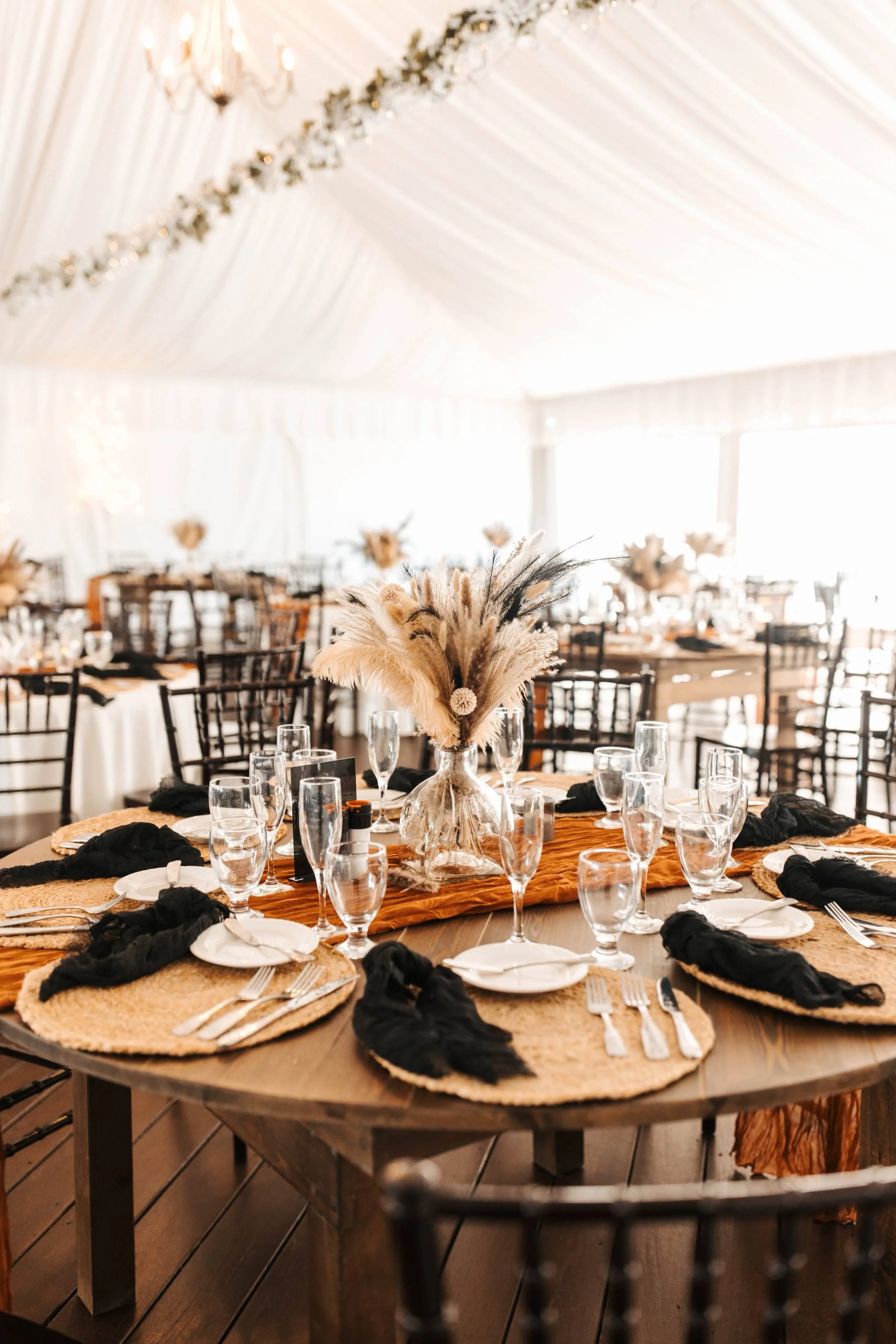 A round dining table decorated with a floral centerpiece featuring feathers and dried flowers, set with glassware, plates, and utensils on woven placemats with black napkins, in a warmly lit banquet hall with draped white ceiling and gold accents.