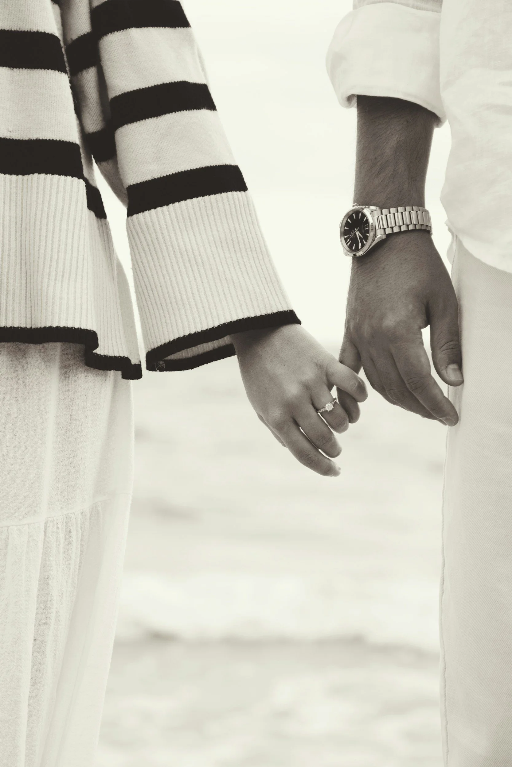 Close-up of a man and woman holding hands, with the woman wearing a striped sweater and the man wearing a watch and a light shirt.