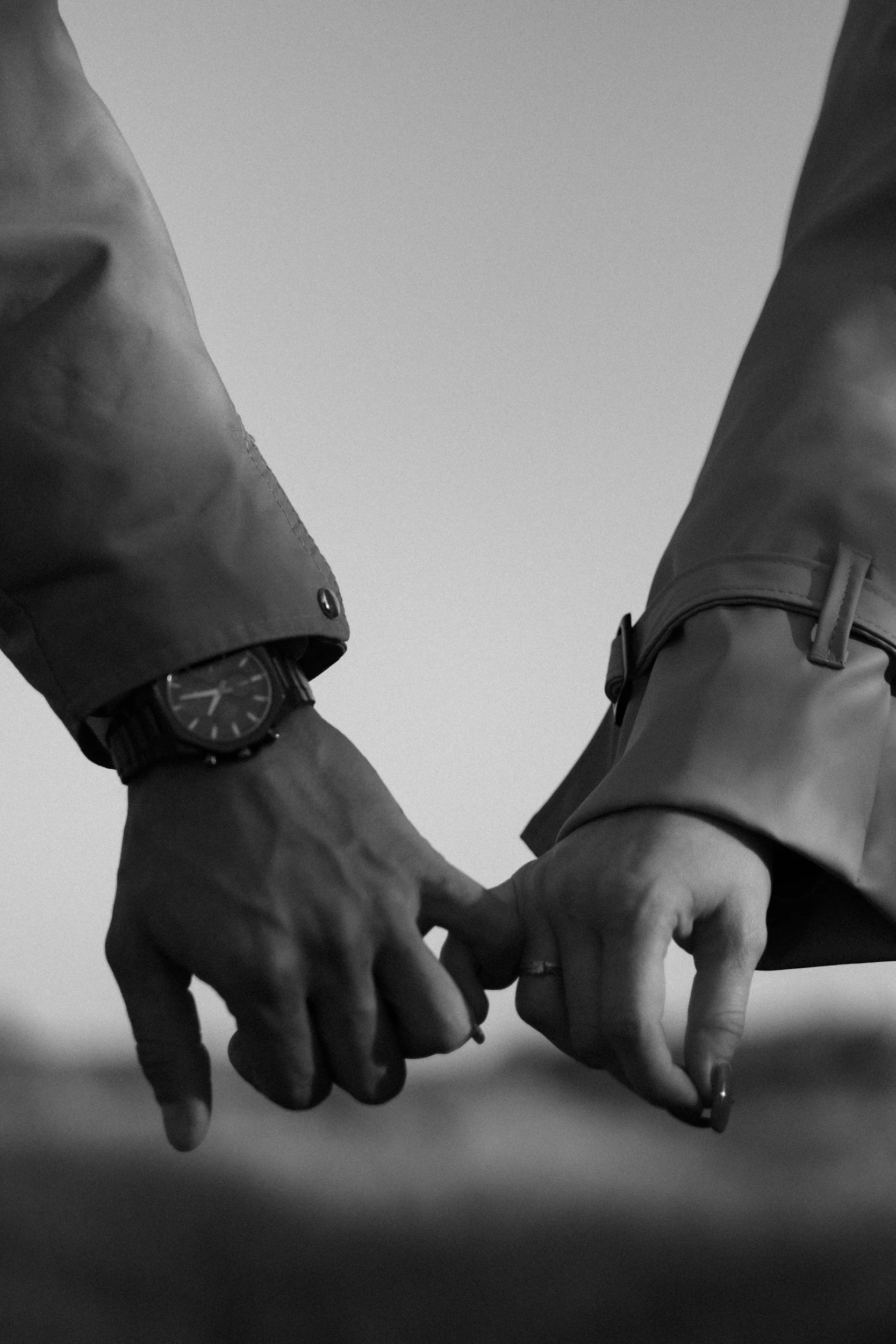 Close-up of two people holding hands, both wearing long-sleeved shirts and watches, with a plain background.