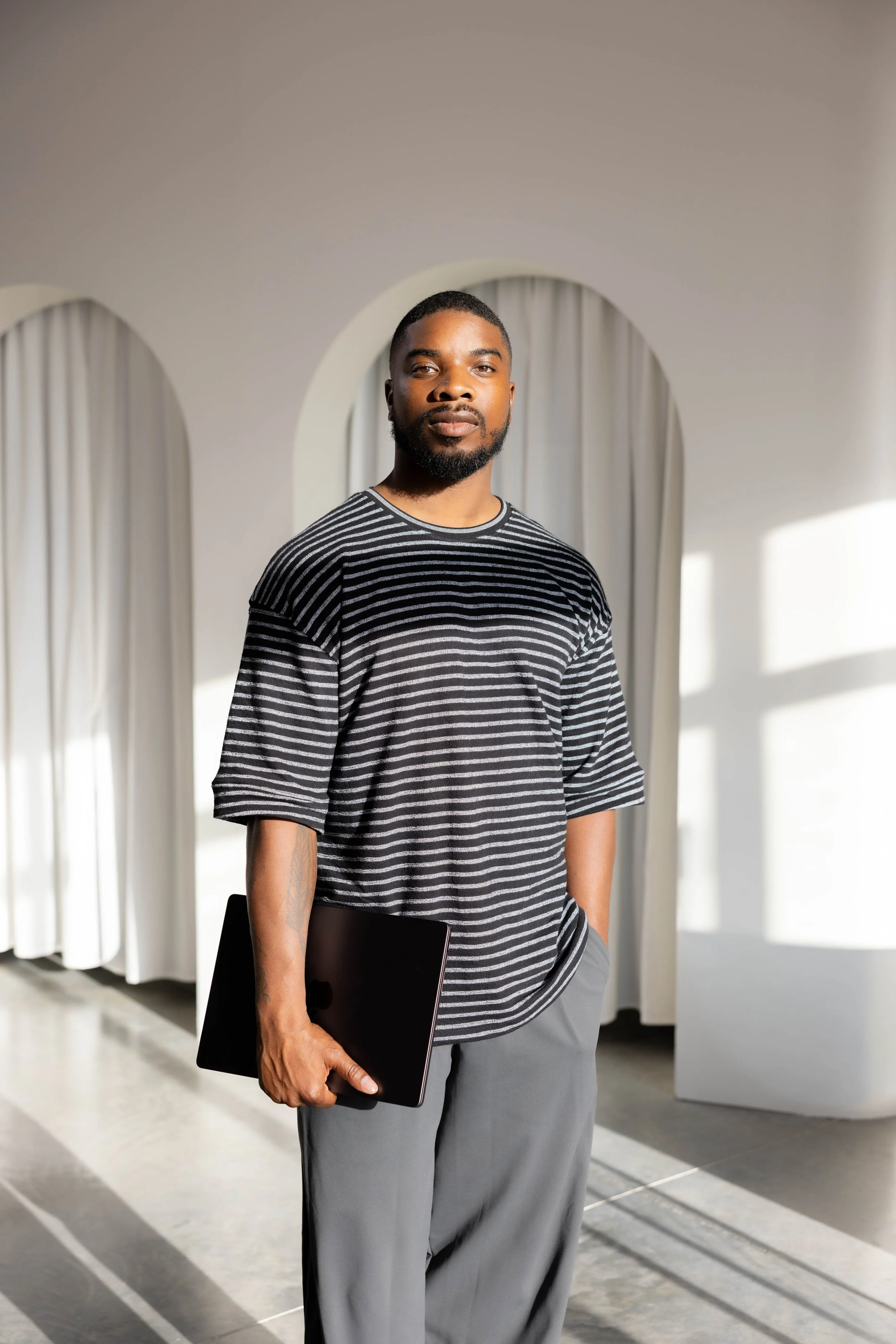 A man with a beard and short hair stands indoors in front of white curtains, holding a black laptop in his right hand, wearing a black and white striped T-shirt and gray pants, with sunlight streaming in Fort Lauderdale, Florida.