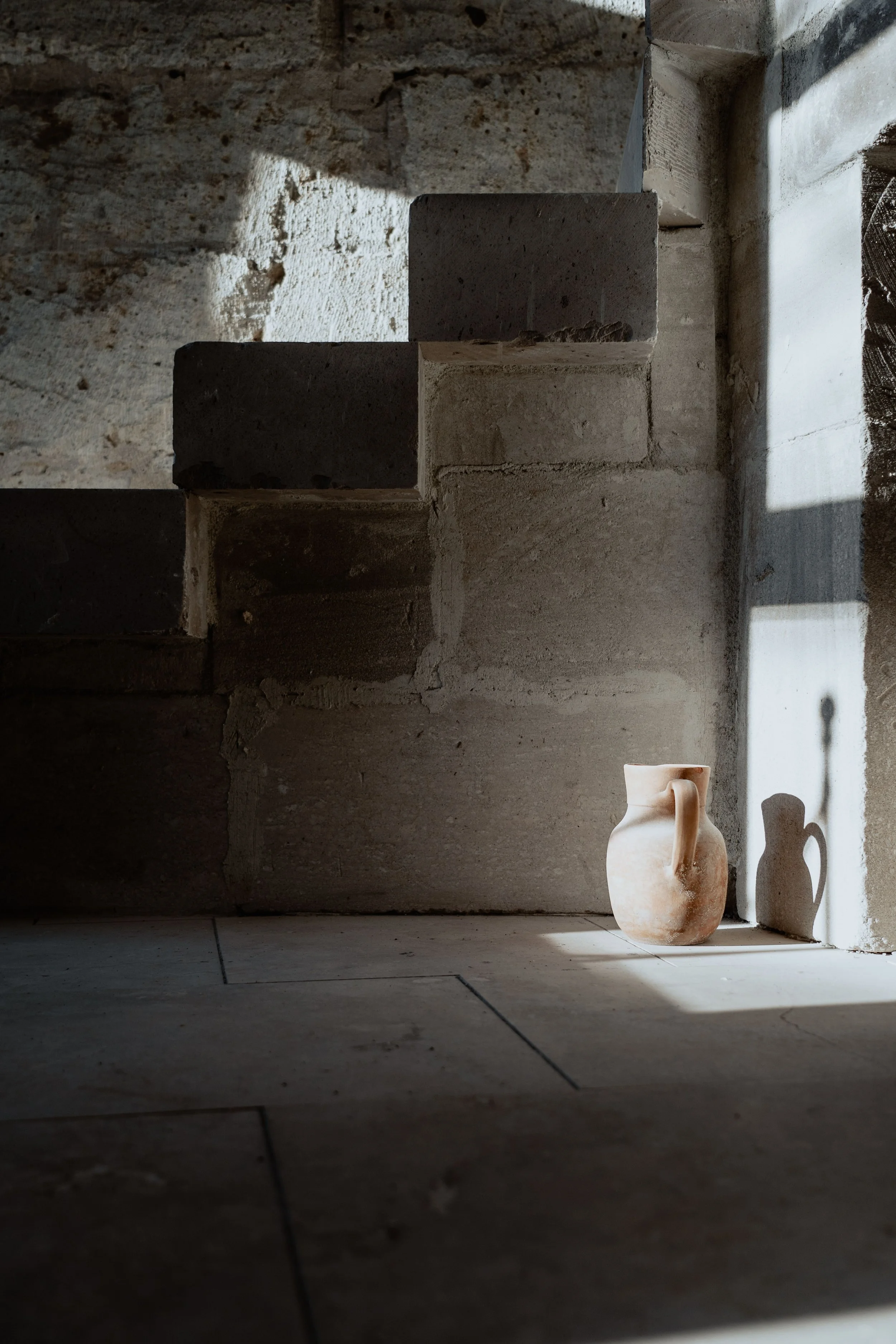 Small ceramic vase with a handle on a tiled floor, with warm natural light casting soft shadows against a textured stone wall in a calming therapy space.