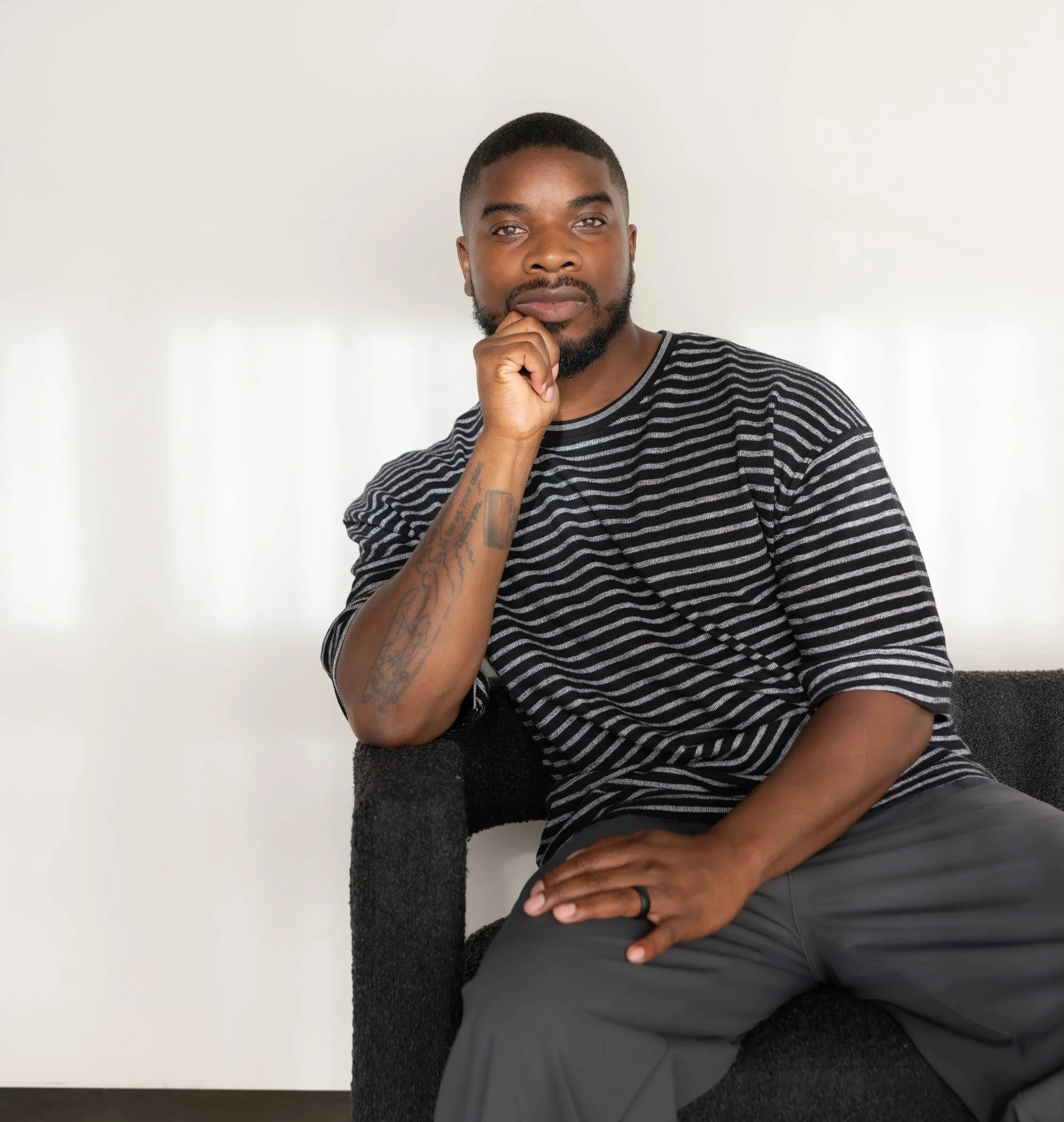 A young man with short hair and a beard, wearing a striped black and gray T-shirt, sitting against a white wall. He has a thoughtful expression, resting his chin on his hand, at a adult ADHD therapy office in Fort Lauderdale, Florida.