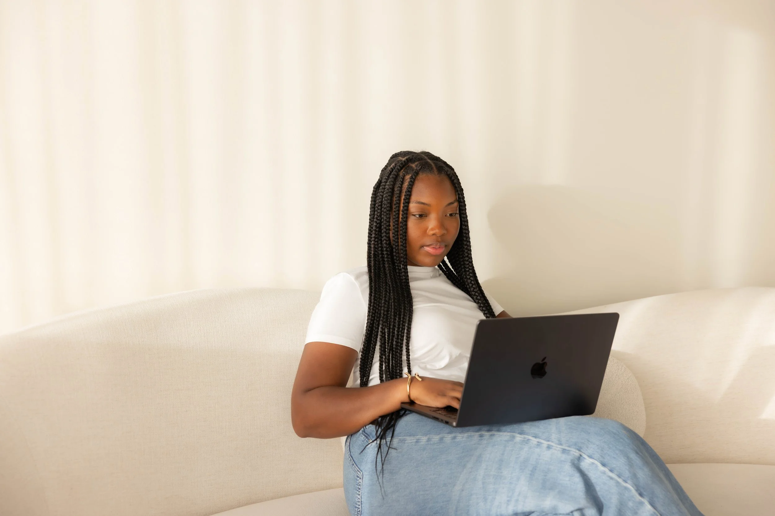 Young woman with braided hair sitting on a beige sofa, working on a laptop with a neutral expression.