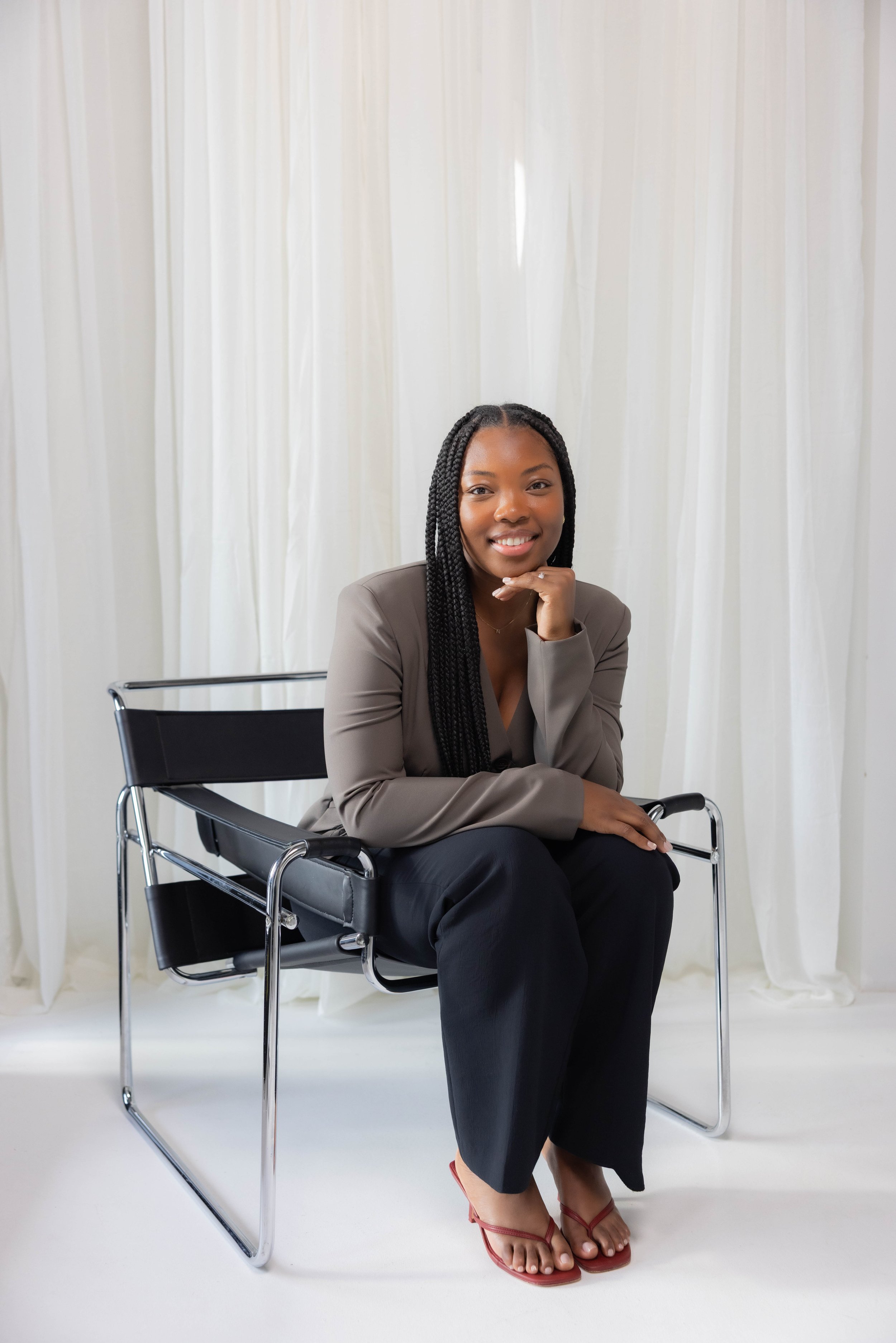 A woman with sitting on an office chair, smiling, with her hand resting on her chin. She is wearing a beige blazer, black pants, and red sandals. She is a individual and couples therapist in Fort Lauderdale, Florida.