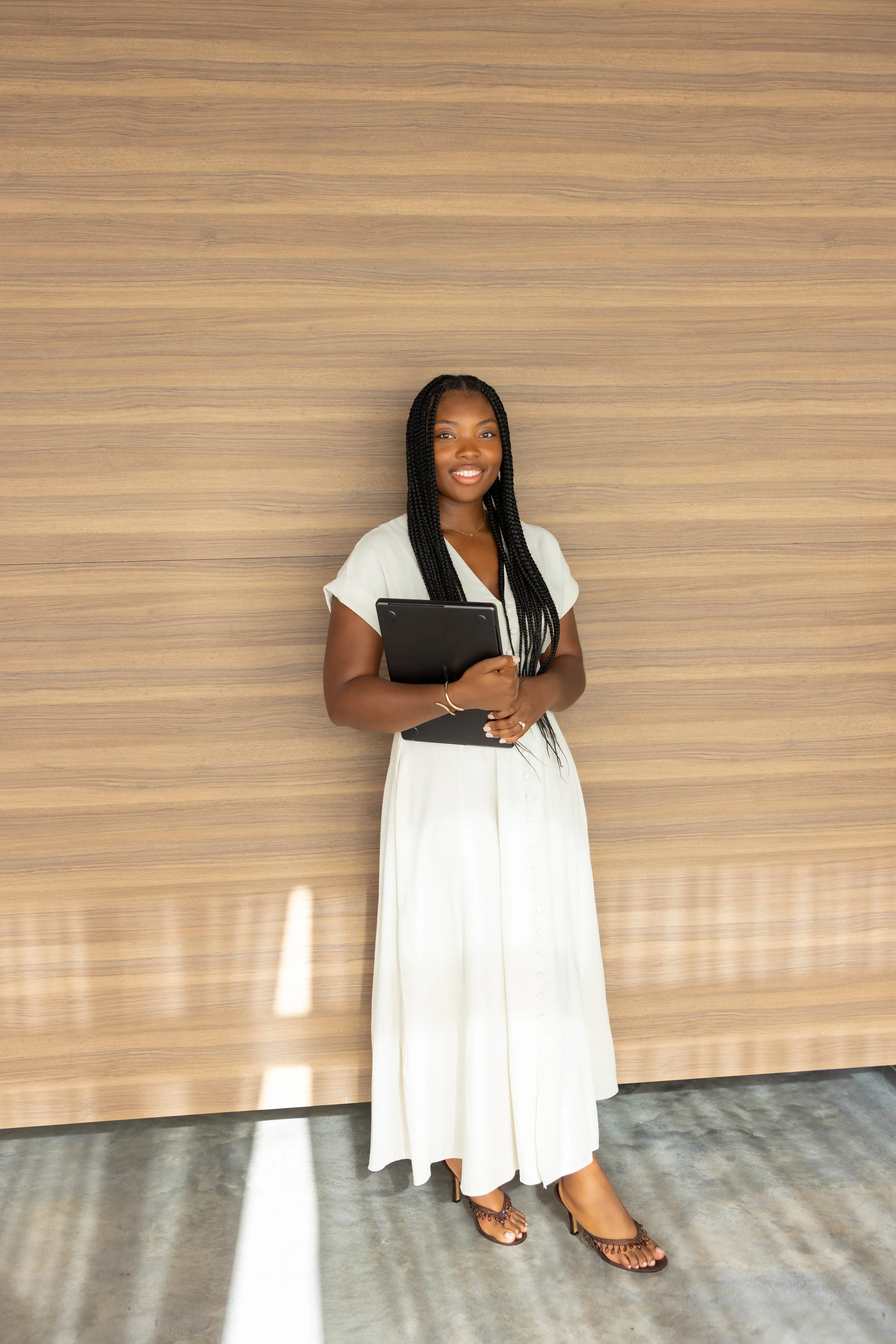 A woman with longhair, wearing a white dress, holding a tablet and smiling, standing in front of a wooden wall. She is a trauma therapist in Fort Lauderdale, Florida.