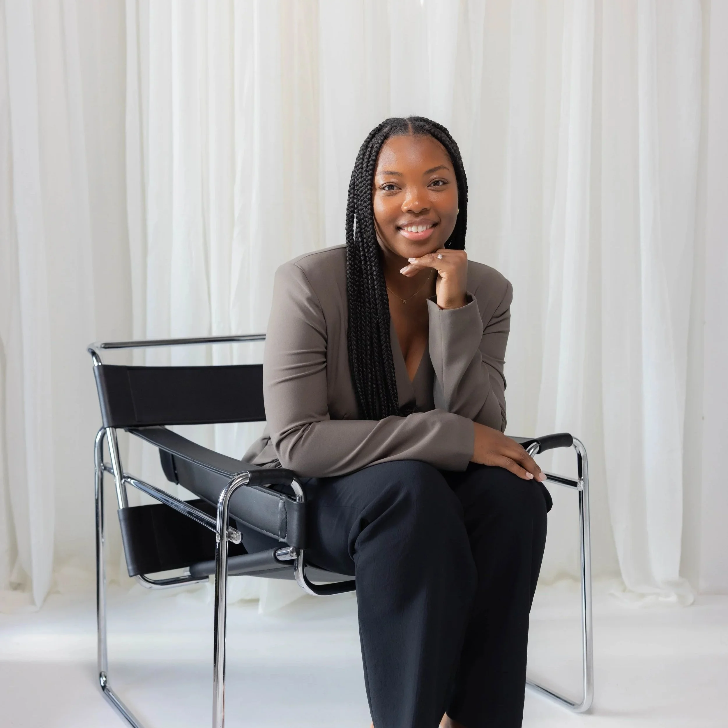 A woman with long braided hair smiling and posing in front of a white curtain, sitting on a black chair with metal armrests, wearing a gray blazer and black pants.