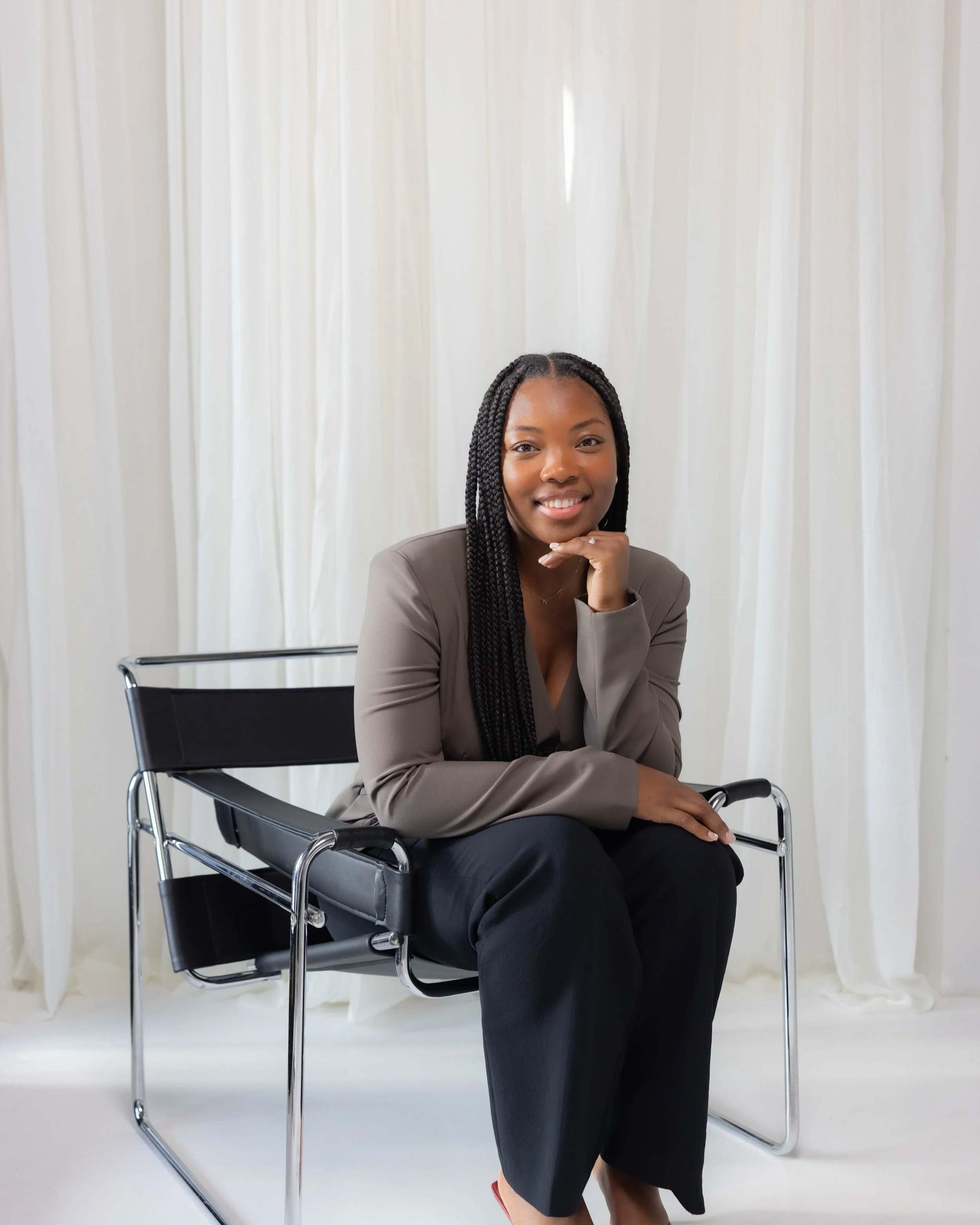 A woman with braided hair wearing a gray blazer and black skirt sitting in a black chair with a metal frame, smiling and resting her chin on her hand, in front of white curtains.