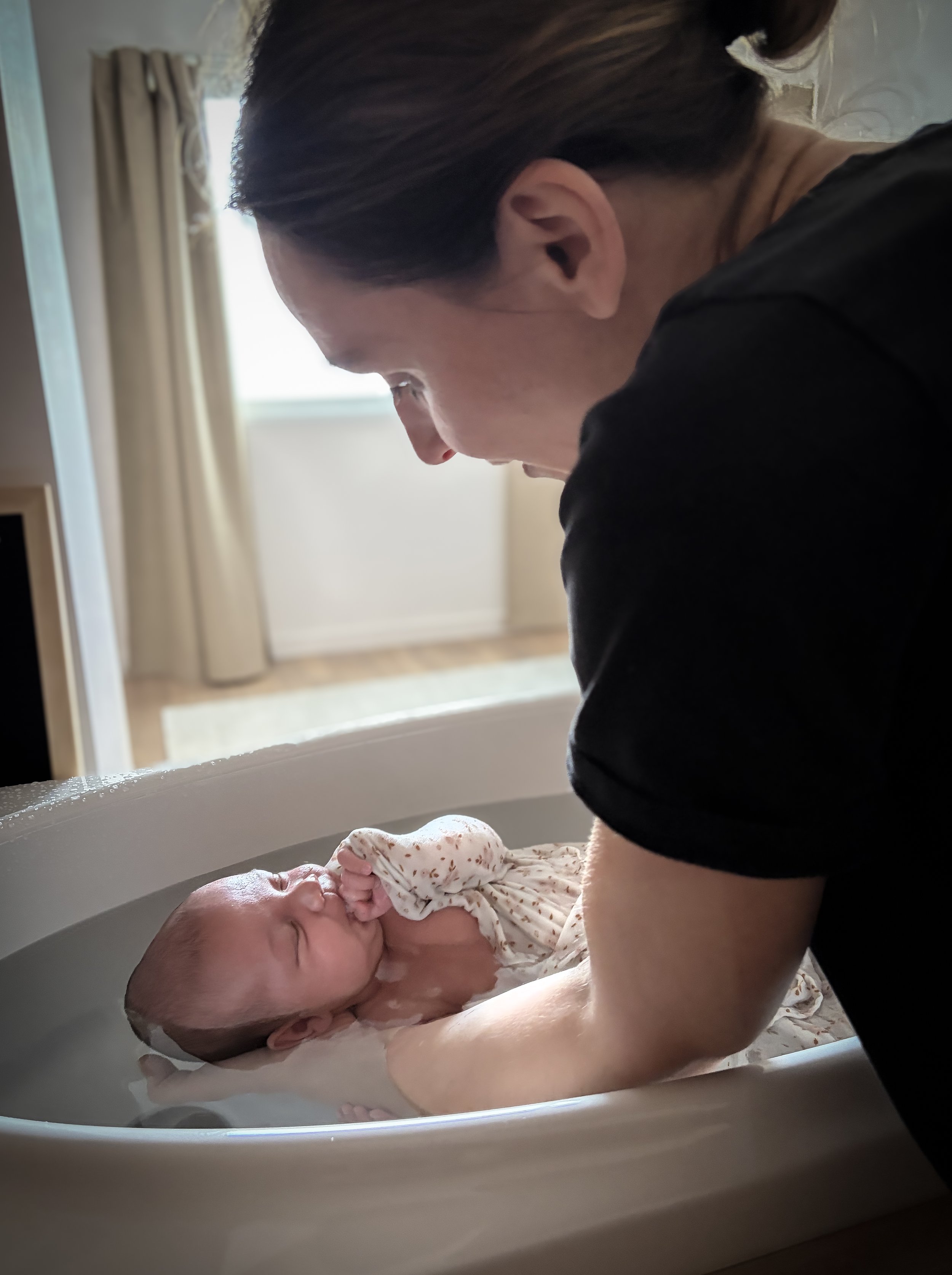 Une femme donne un bain à un nouveau-né dans une baignoire, l'enfant couvre son visage avec une main.