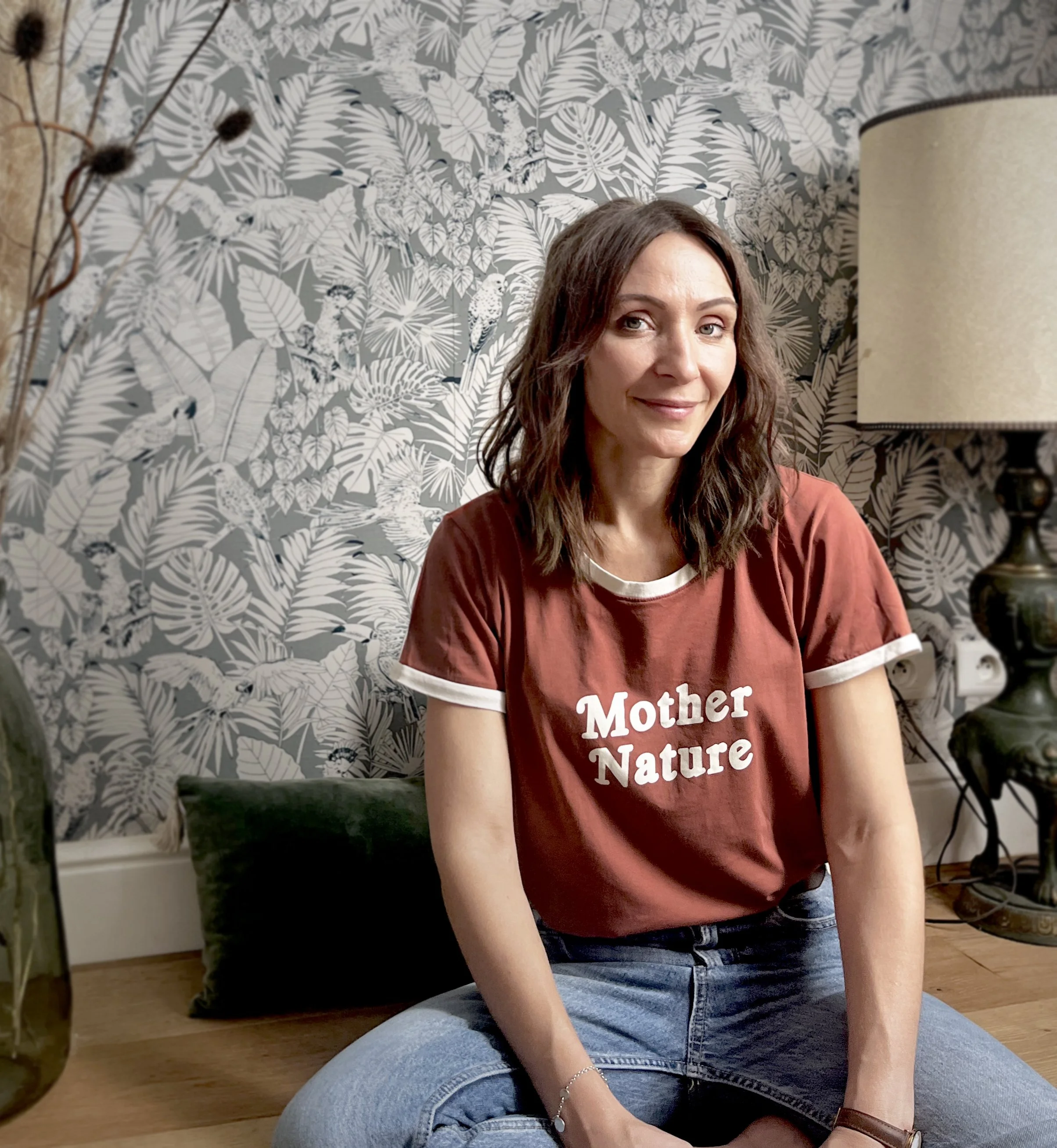 Une femme assise par terre, portant un t-shirt rouge avec le texte "Mother Nature", dans un intérieur avec un papier peint à motif de feuilles et d'oiseaux. Elle sourit légèrement, les cheveux marrons ondulés.