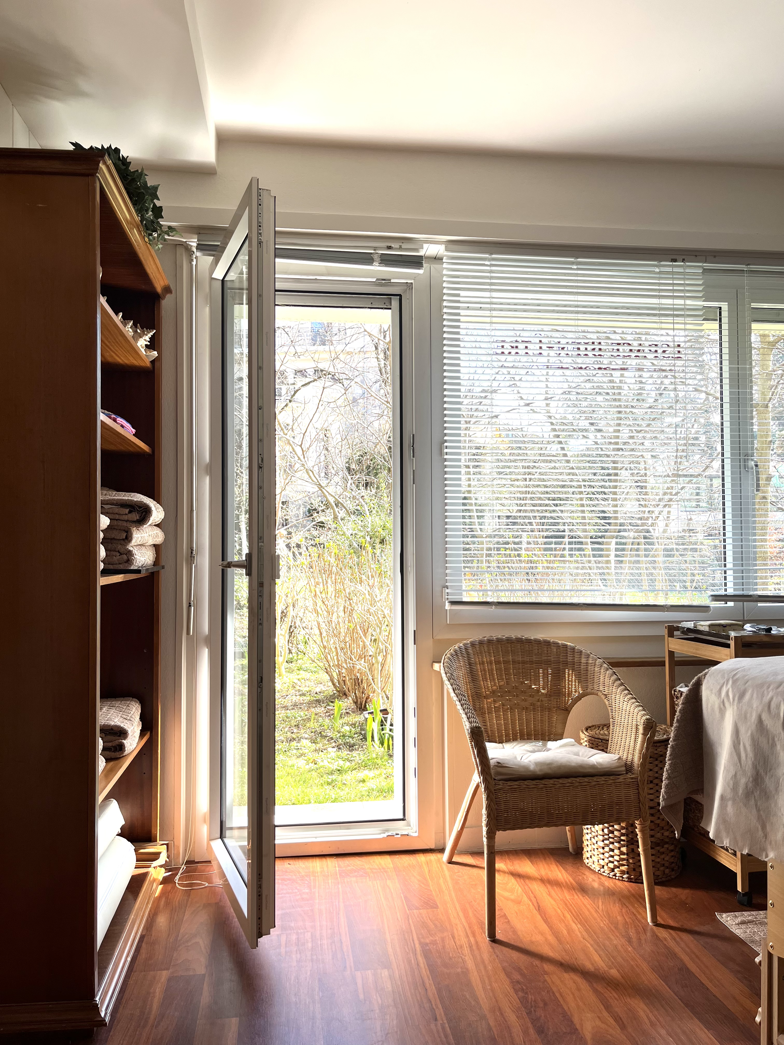 Une pièce lumineuse avec un sol en bois foncé, une fenêtre avec volets horizontaux laissant entrer la lumière et un jardin visible à l'extérieur. Il y a une chaise en rotin avec coussin, une table en bois à côté du lit, et une étagère en bois avec des serviettes ou des draps pliés. La pièce donne une ambiance chaleureuse et accueillante.