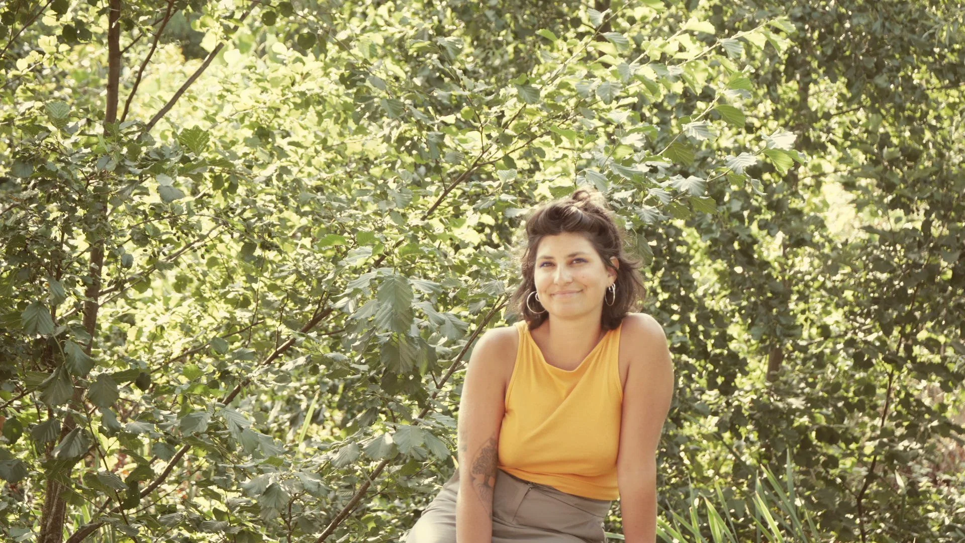 Jeune femme souriante portant un haut jaune assise parmi des arbres en plein air, soleil brillant, ambiance naturelle et détendue.