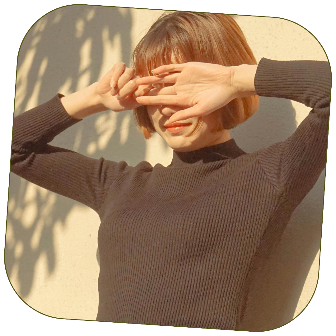 A woman with short, reddish-brown hair wearing a dark ribbed turtleneck sweater, standing outside against a textured wall, shielding her eyes with her right hand while smiling.