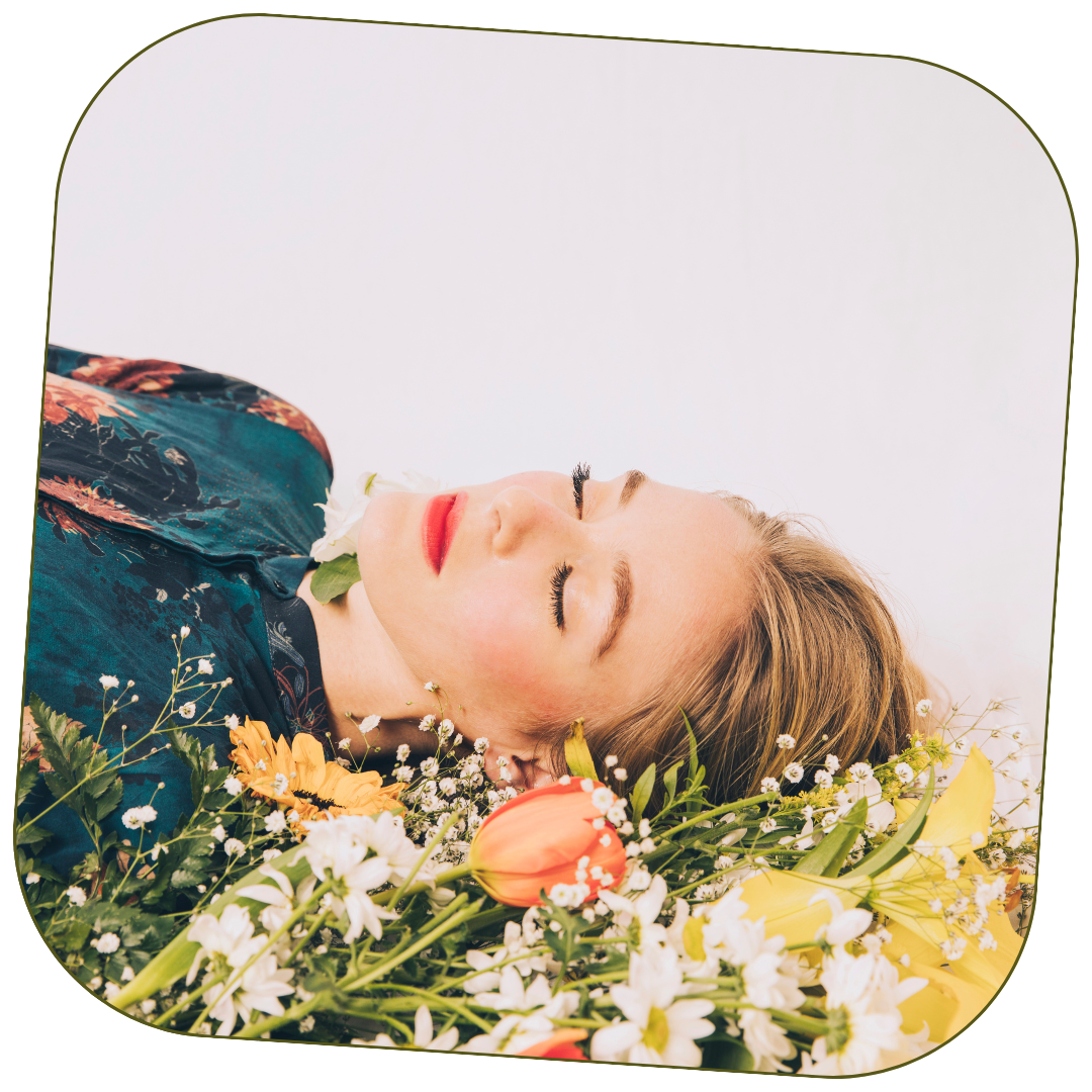 A woman lying on her back with closed eyes, surrounded by a variety of colorful flowers including orange tulips, white daisies, and yellow lilies, on a plain white background.