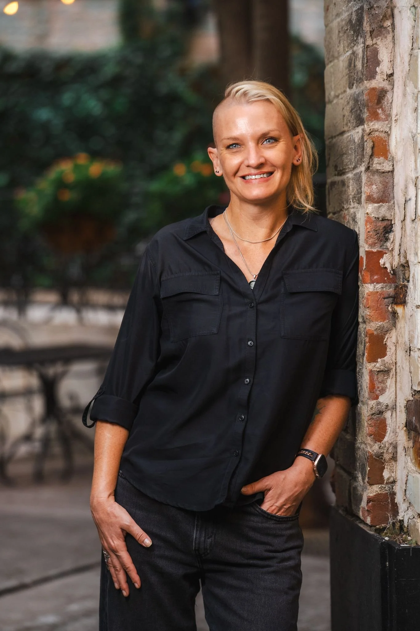 A woman with short, asymmetrical blonde hair, wearing a black button-up shirt with rolled-up sleeves, standing outdoors against a brick wall, with a blurred background of green foliage.