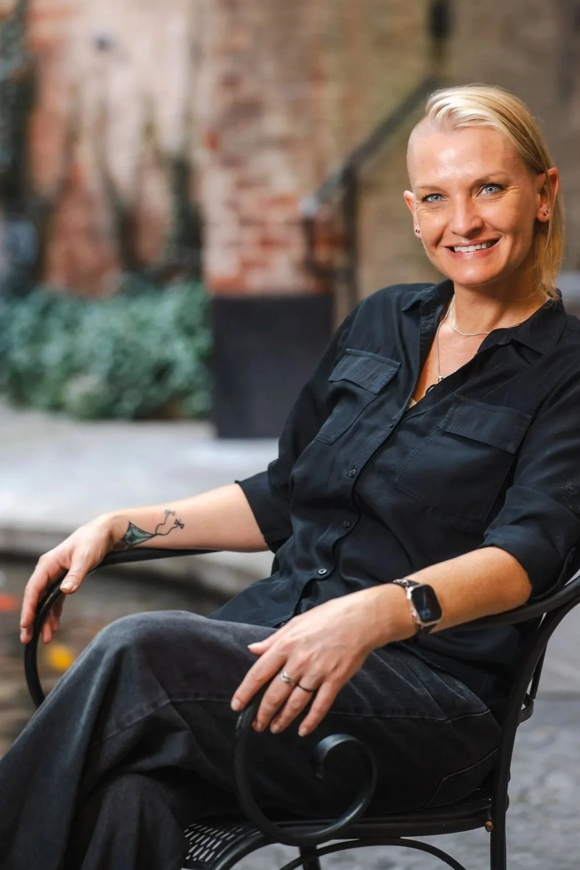 A woman with short blonde hair, wearing a black shirt, smiling while sitting in a black chair outdoors near a brick wall and some plants.