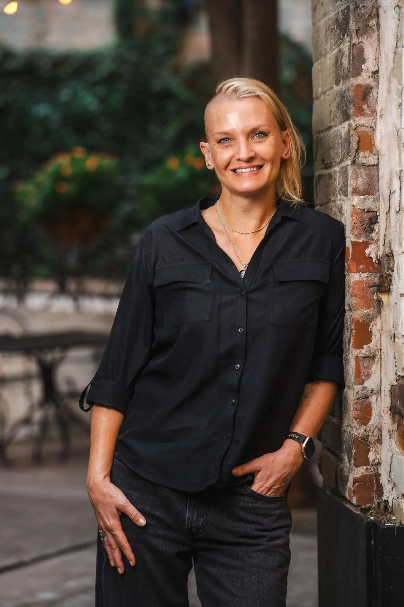 A woman with short blonde hair, wearing a black shirt and black pants, standing outdoors against a brick wall and lush greenery in the background.