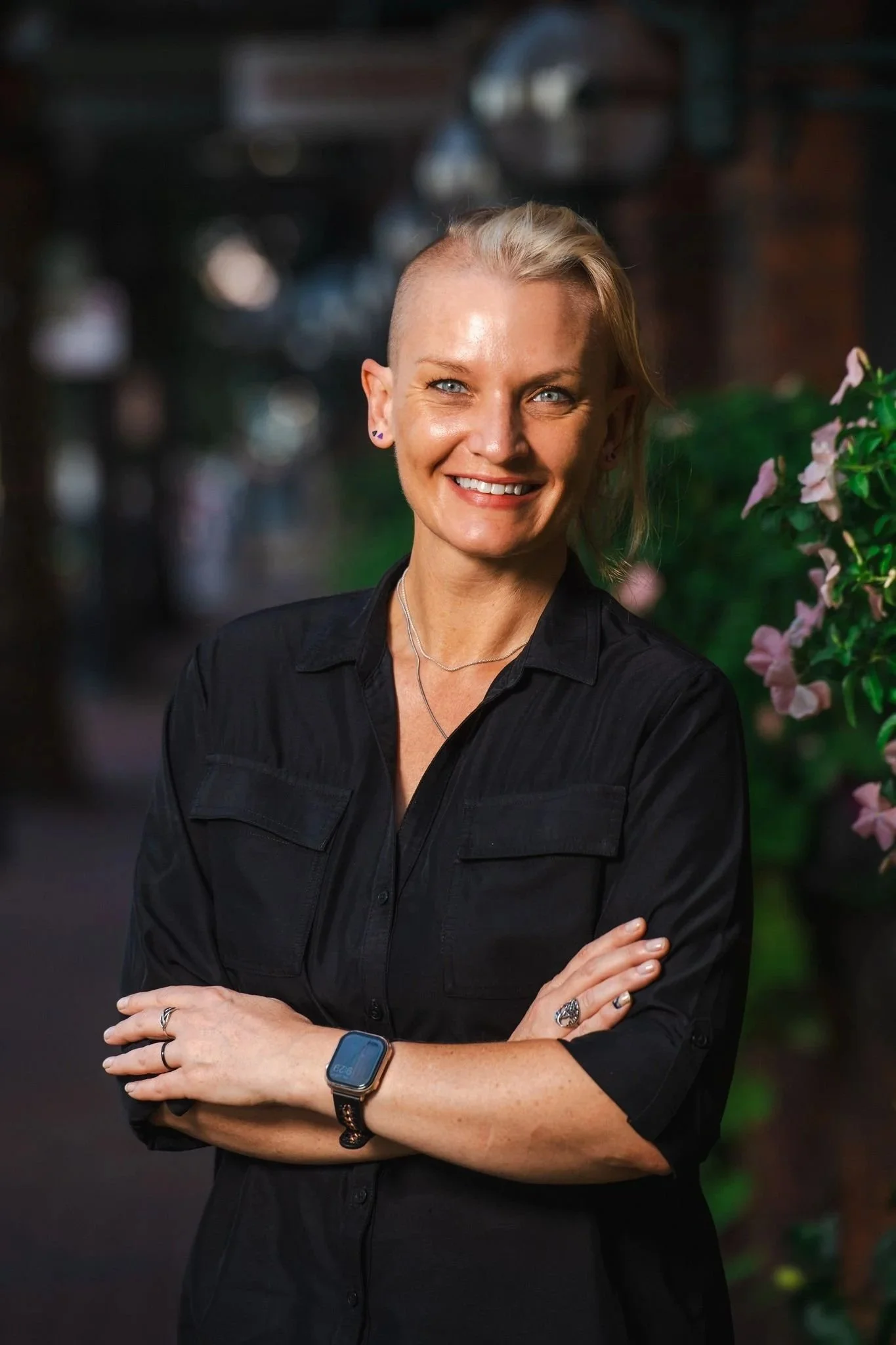 Portrait of a smiling woman with short bleach blonde hair and blue eyes, wearing a black button-up shirt, wristwatch, rings, and earrings, with arms crossed in a dark, blurry background with purple flowers on the right.