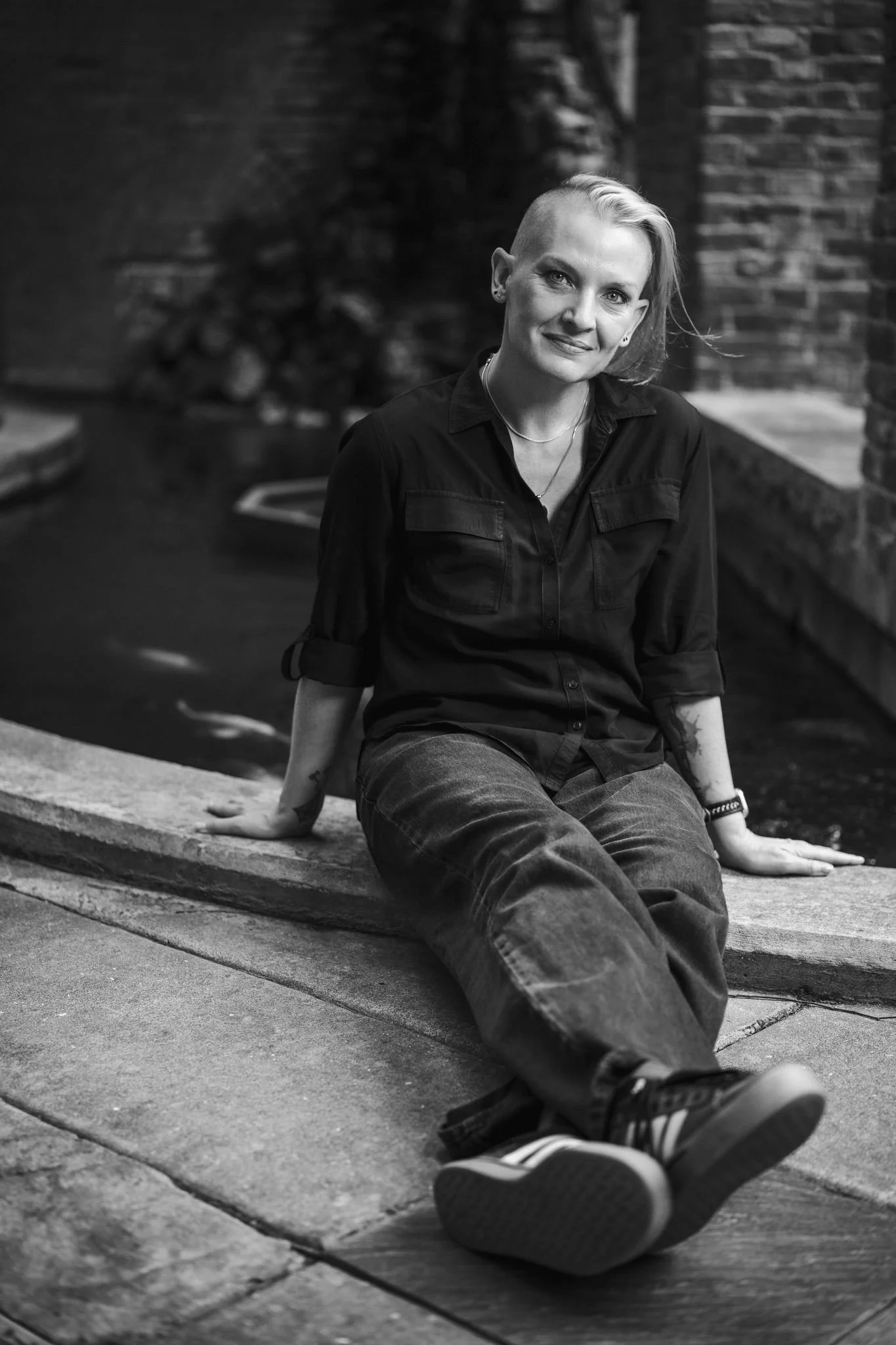 A woman with short blonde hair, wearing a black shirt and jeans, is sitting on the sidewalk with her back against a brick wall. She is smiling and looking at the camera on a street or outdoor urban setting.