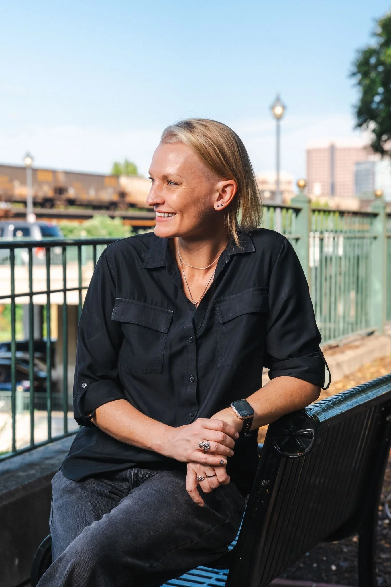 A smiling woman with short blonde hair and pierced ears sitting on a black metal bench outdoors, wearing a black shirt and a smartwatch, with a cityscape and train in the background on a sunny day.
