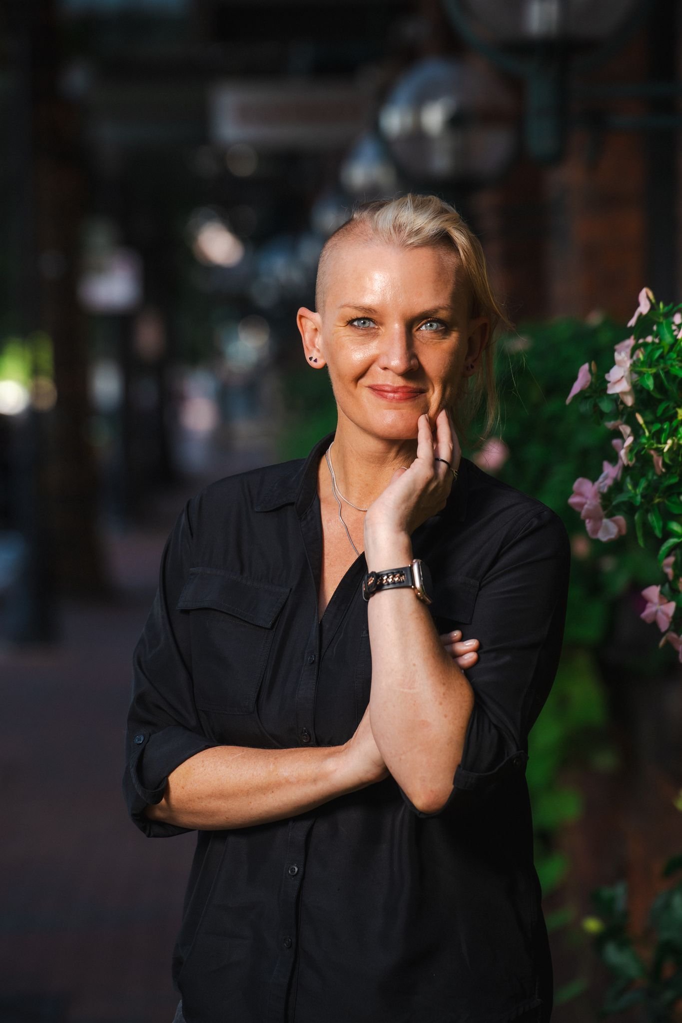 A woman with short, blonde, partially shaved hair, blue eyes, and light skin, posing outdoors with lush greenery and pink flowers nearby, smiling softly while resting her hand on her chin.