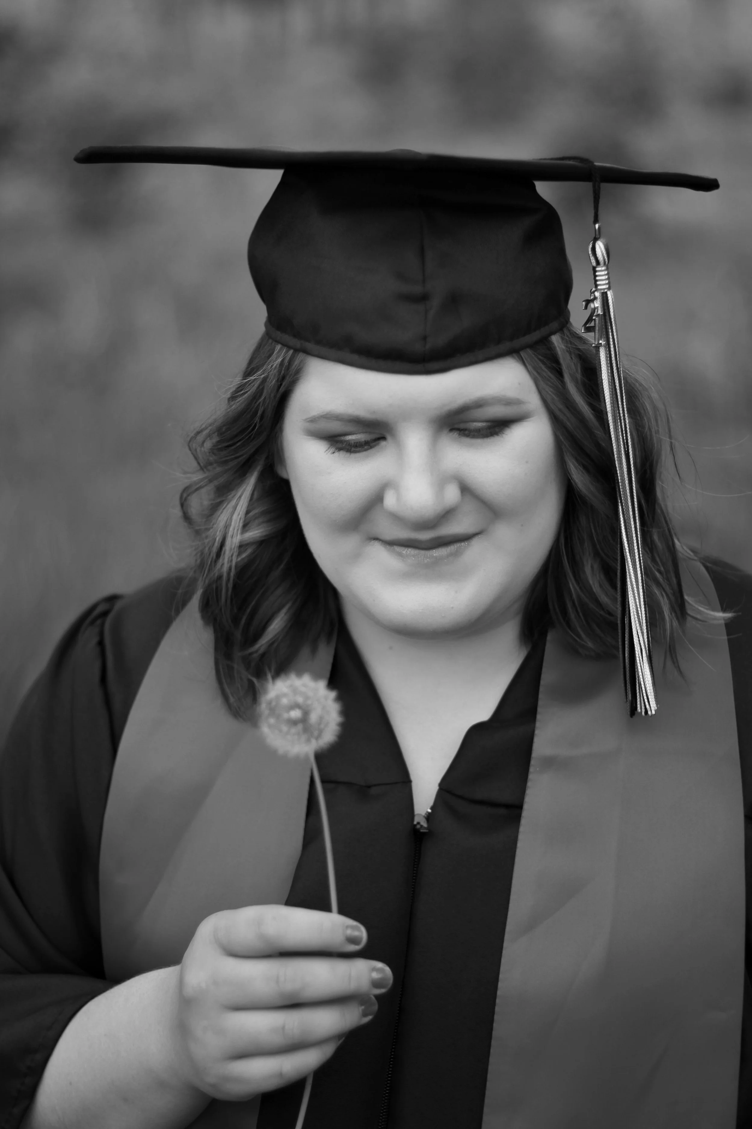 A woman in a graduation gown and cap is looking at a dandelion, which she is holding with her left hand. The photo is in black and white.