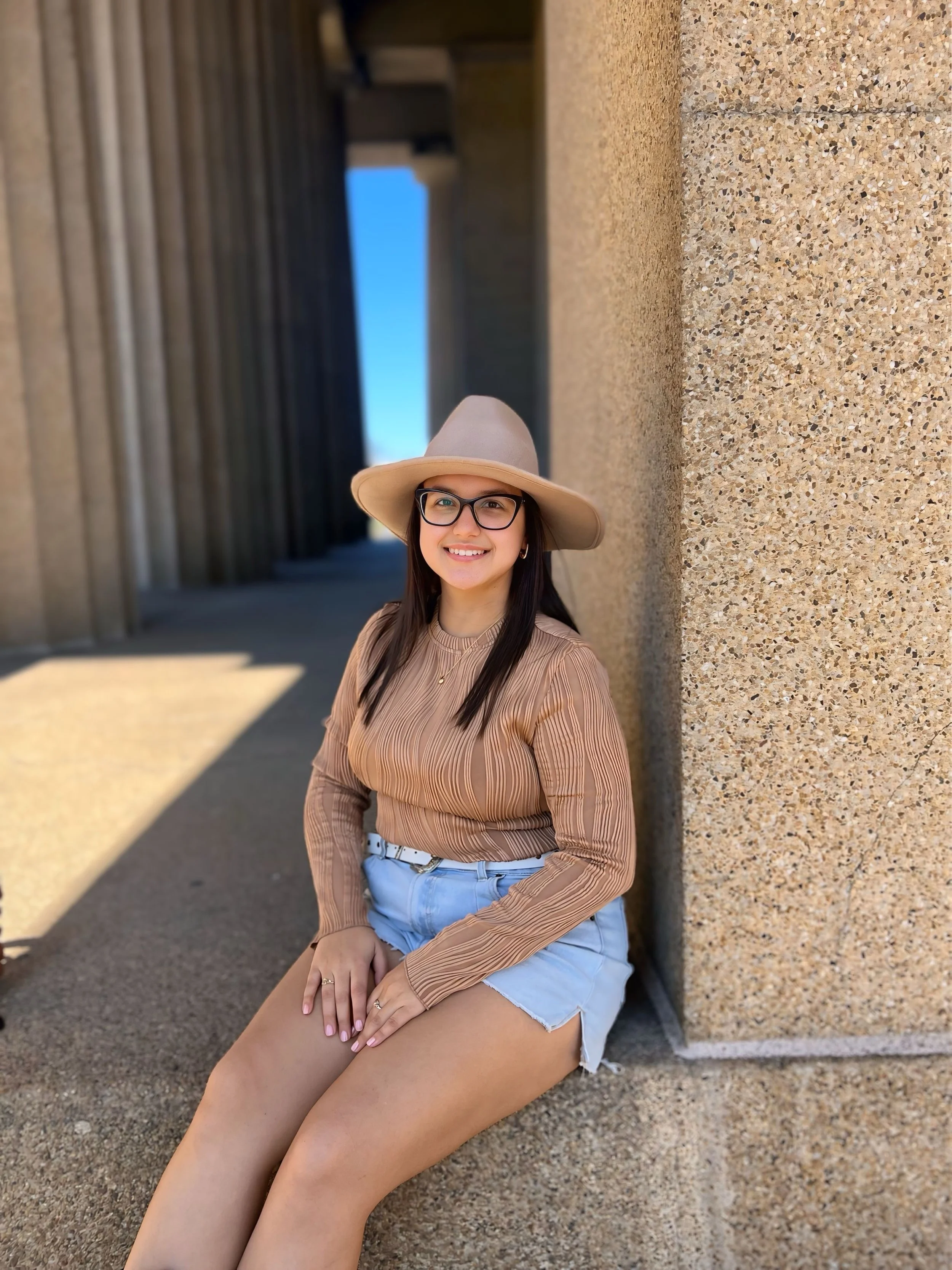 A woman sitting outdoors against a textured concrete wall, smiling, wearing a wide-brimmed beige hat, glasses, a brown striped long-sleeve top, and light blue denim shorts.