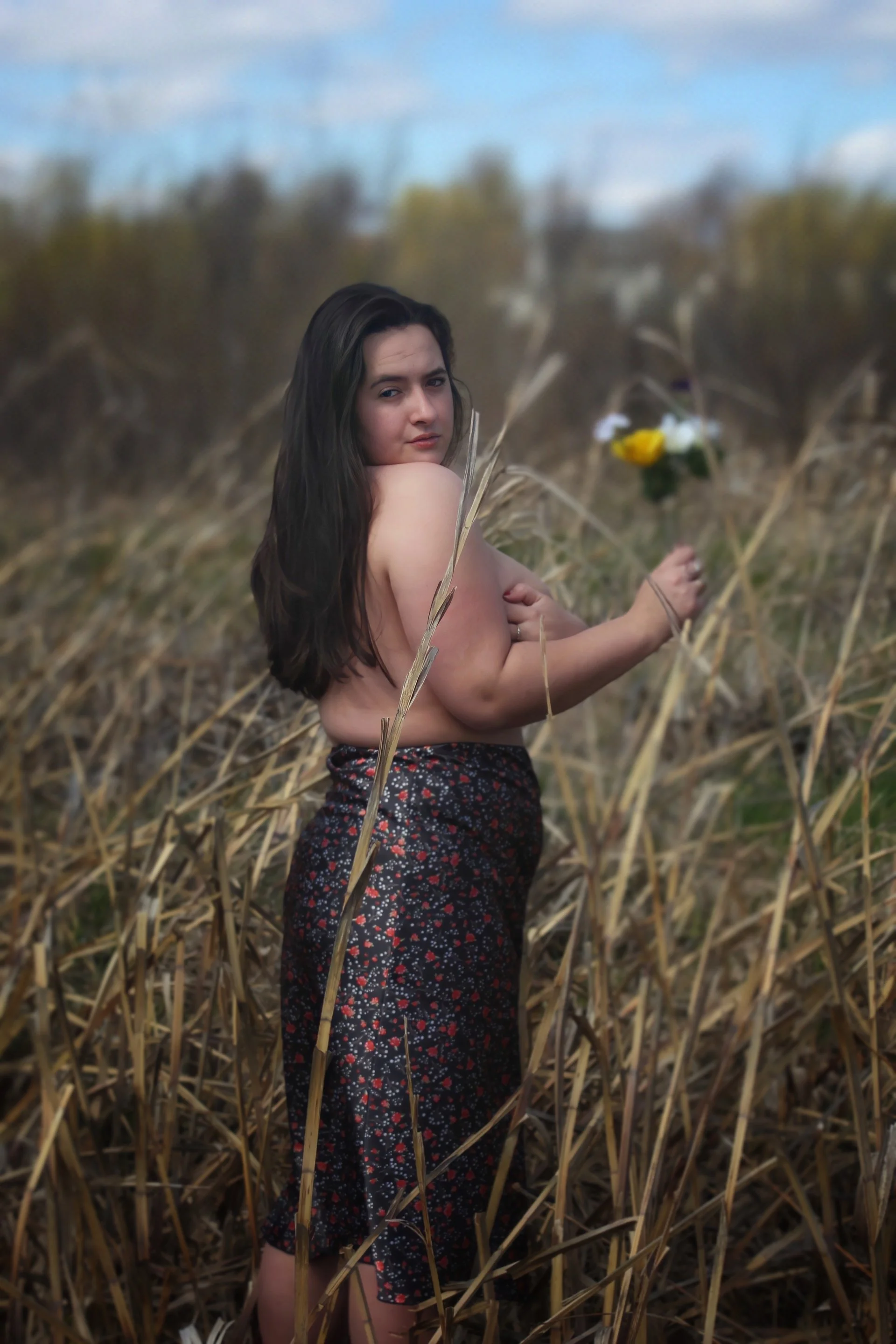 A woman with long dark hair standing topless in a field of dry tall grass, holding a yellow and white flower, with a blurred background of trees and a sky with some clouds.