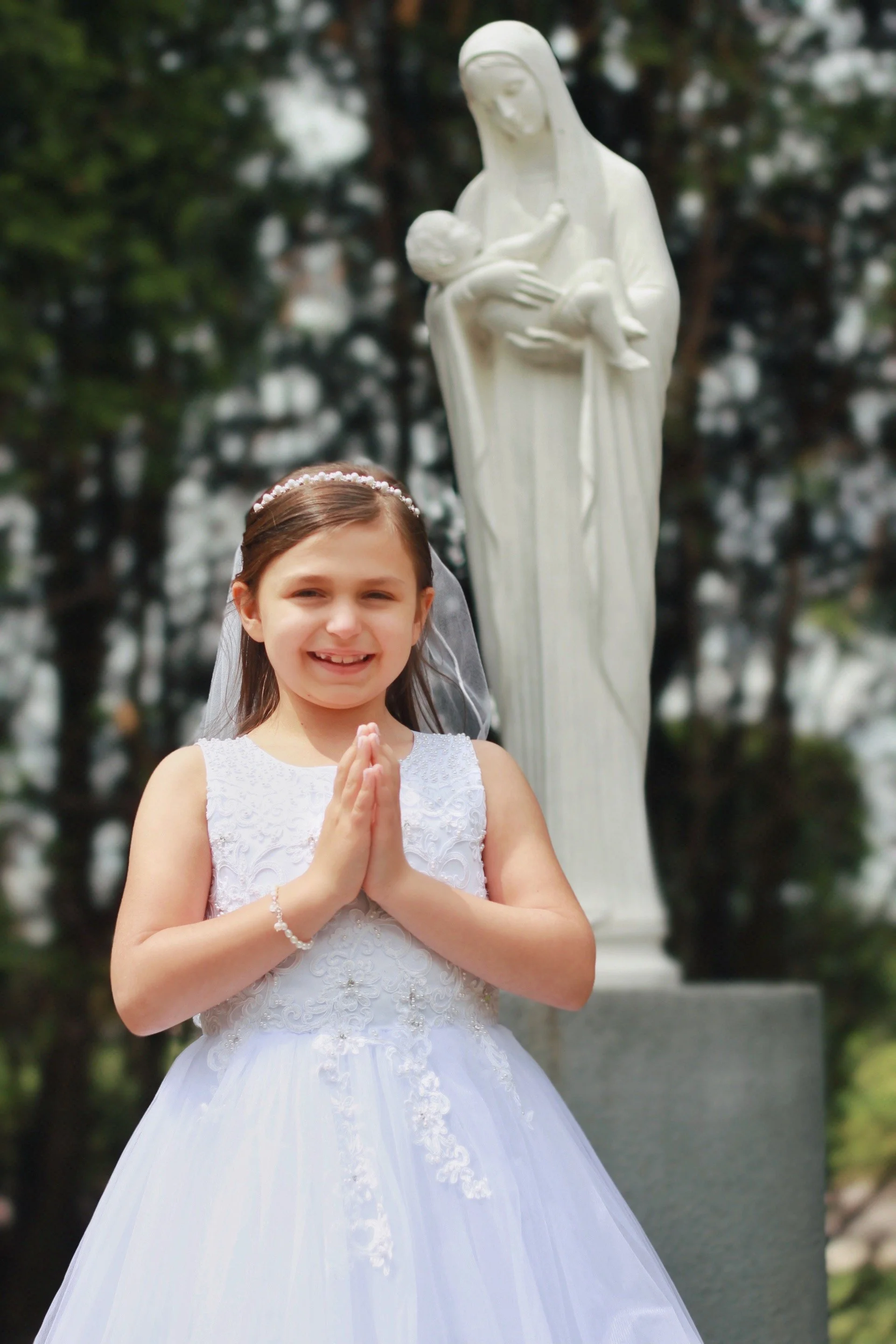 A young girl in a white lace dress and veil standing outdoors in front of a statue of Mary holding baby Jesus, with trees in the background.