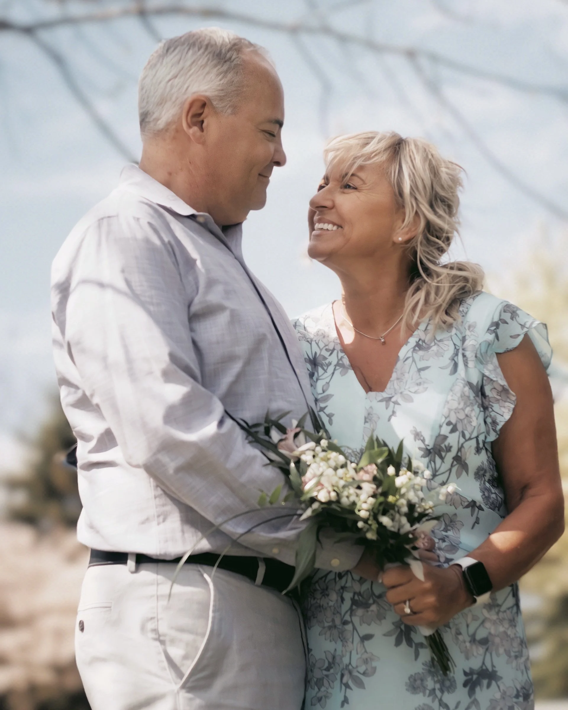 An elderly couple smiling at each other during a wedding ceremony, with the man wearing a light-colored shirt and holding a bouquet of flowers.