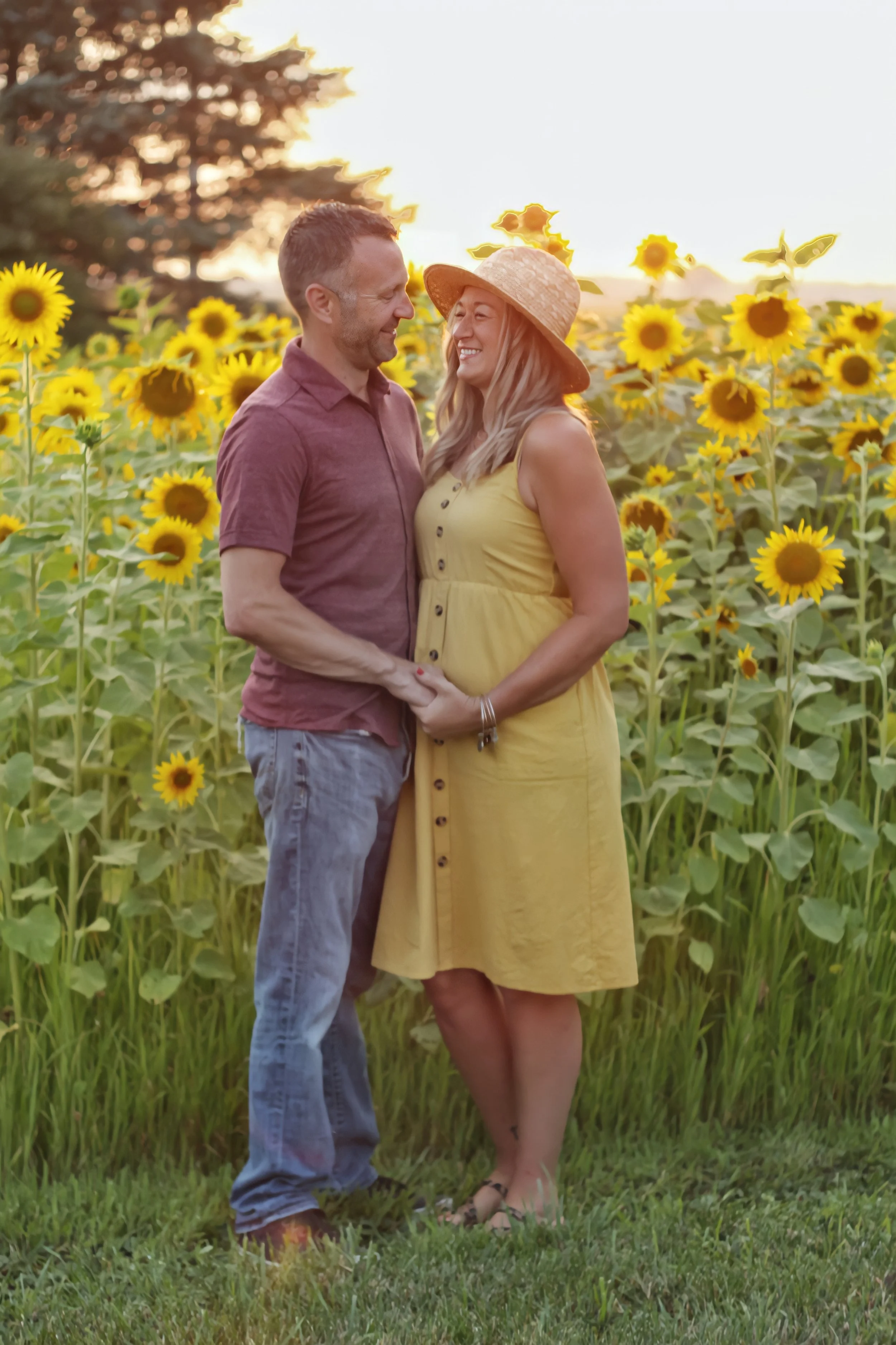 A couple standing in a sunflower field during sunset, smiling at each other. The woman is wearing a yellow dress and a wide-brimmed sun hat, while the man is dressed in a maroon shirt and jeans.