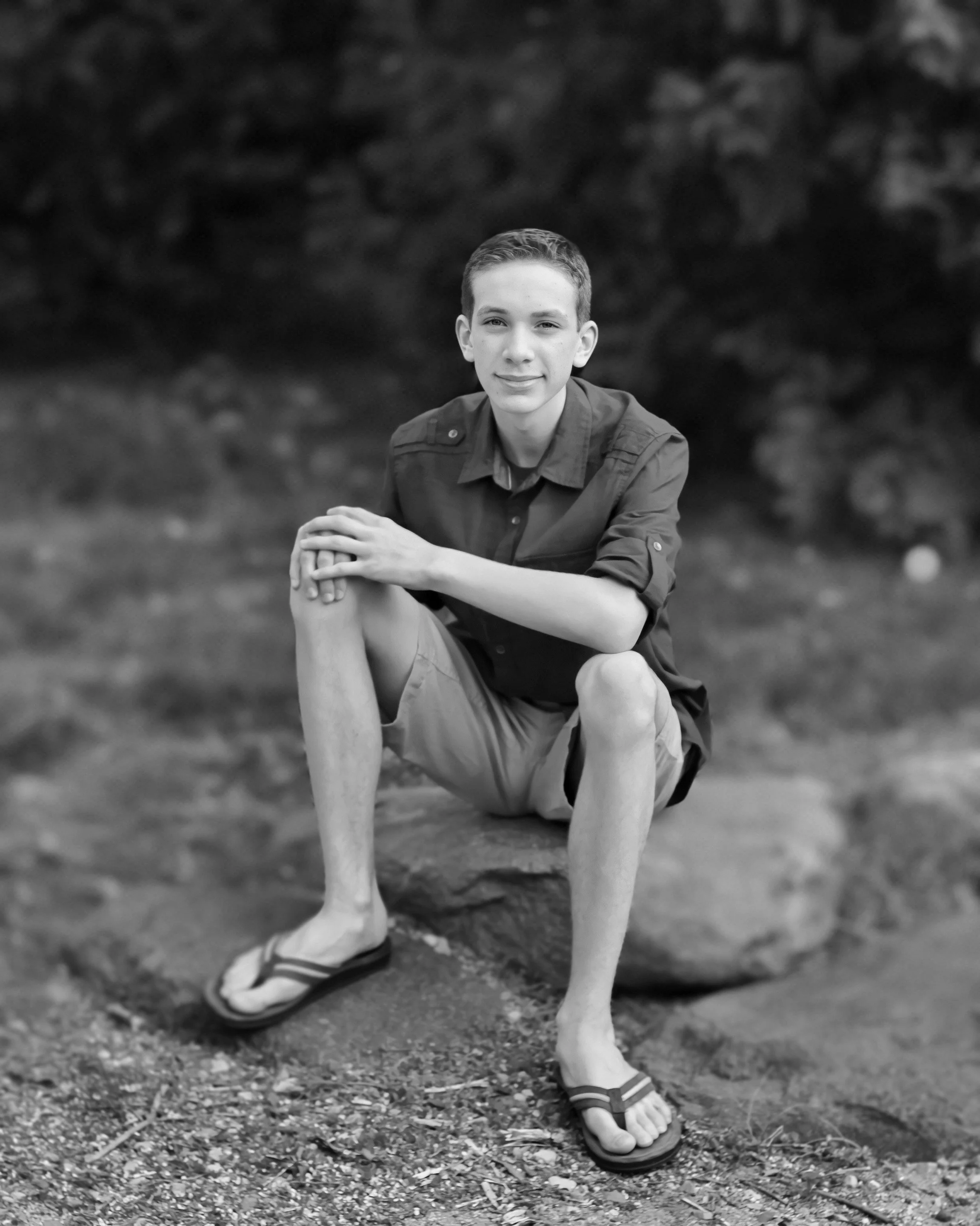 A young man with short hair sitting outdoors with one leg crossed over the other, smiling slightly, wearing a button-up shirt, shorts, and sandals. The black-and-white photo shows his right hand resting on his knee with a background of trees and grass.