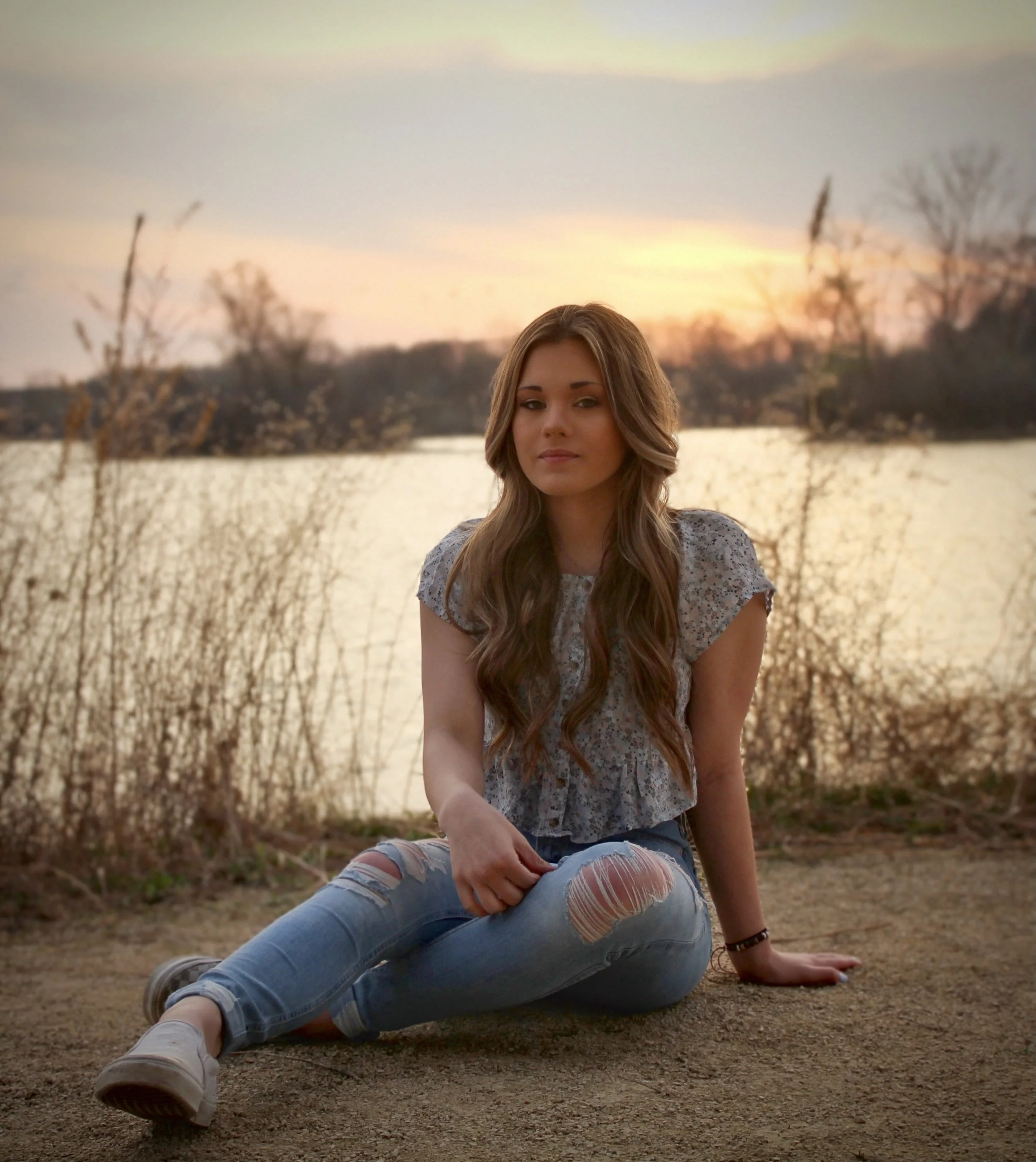A young woman with long brown hair wearing a gray lace top and ripped jeans, sitting on a dirt ground near a lake with trees in the background during sunset.