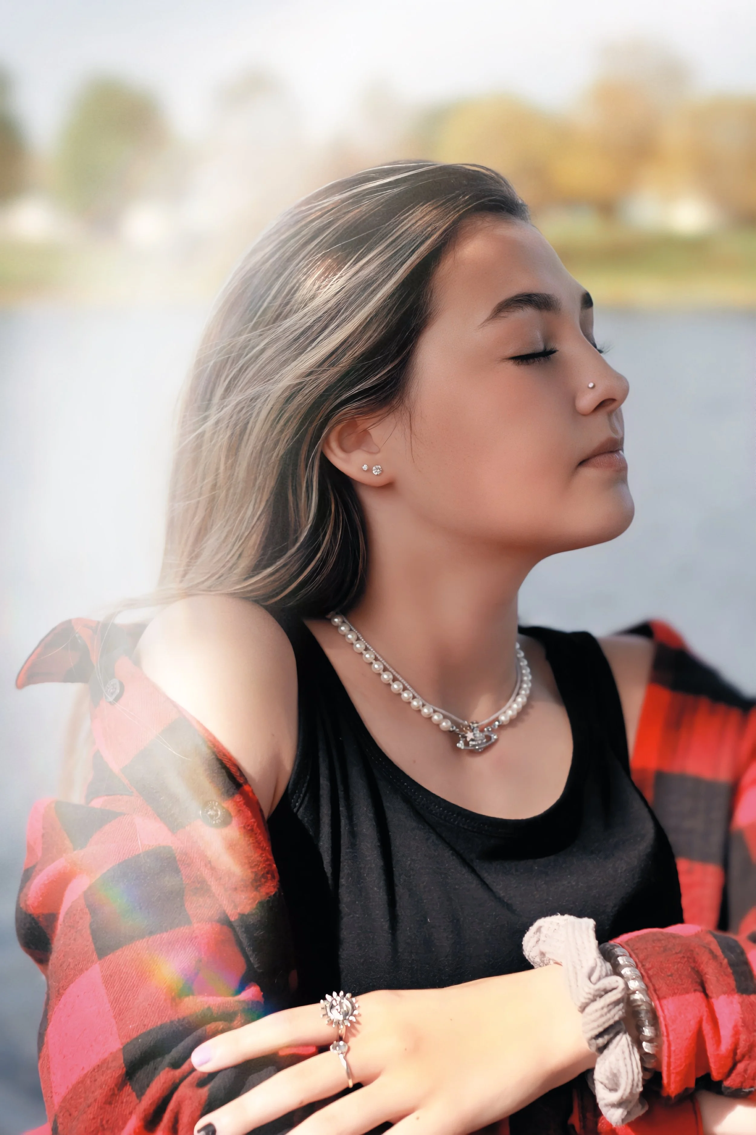A young woman with long blonde hair, closed eyes, and a nose piercing, wearing jewelry including earrings, a necklace, and bracelets, appears to be in a moment of calm or meditation outdoors with a blurred background of trees and water.